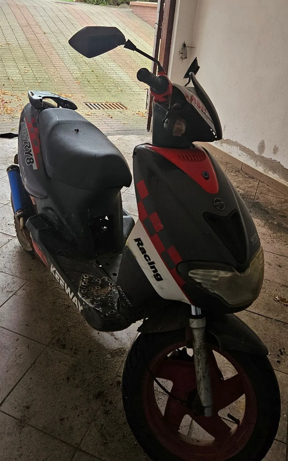 Czarny skuter marki Keeway z czerwonymi akcentami i biało-czerwonymi szachownicami. Na boku widnieją napisy „Racing” oraz „RV8”. Pojazd posiada czerwone felgi i jest zaparkowany na płytkach przed budynkiem.