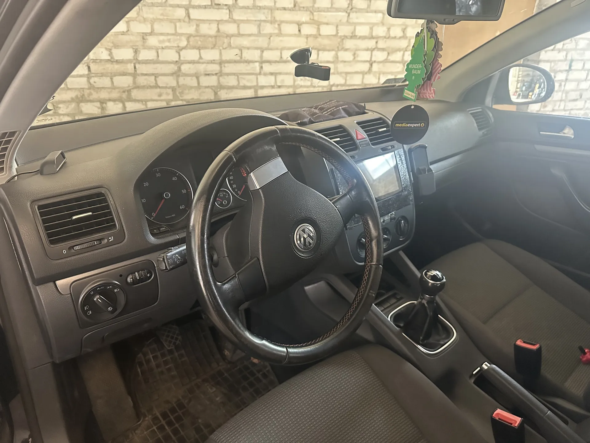 Wnętrze samochodu z kierownicą marki Volkswagen, tablicą rozdzielczą z licznikami prędkości i obrotomierzem, szarymi siedzeniami oraz widoczną przez przednią szybę ceglaną ścianą. Na lusterku wstecznym wiszą aromatyzatory: zielony w kształcie choinki z napisem "WUNDER-BAUM" oraz okrągły czarny z napisem "mediaexpert". W centralnej konsoli znajduje się ekran dotykowy i dźwignia zmiany biegów.