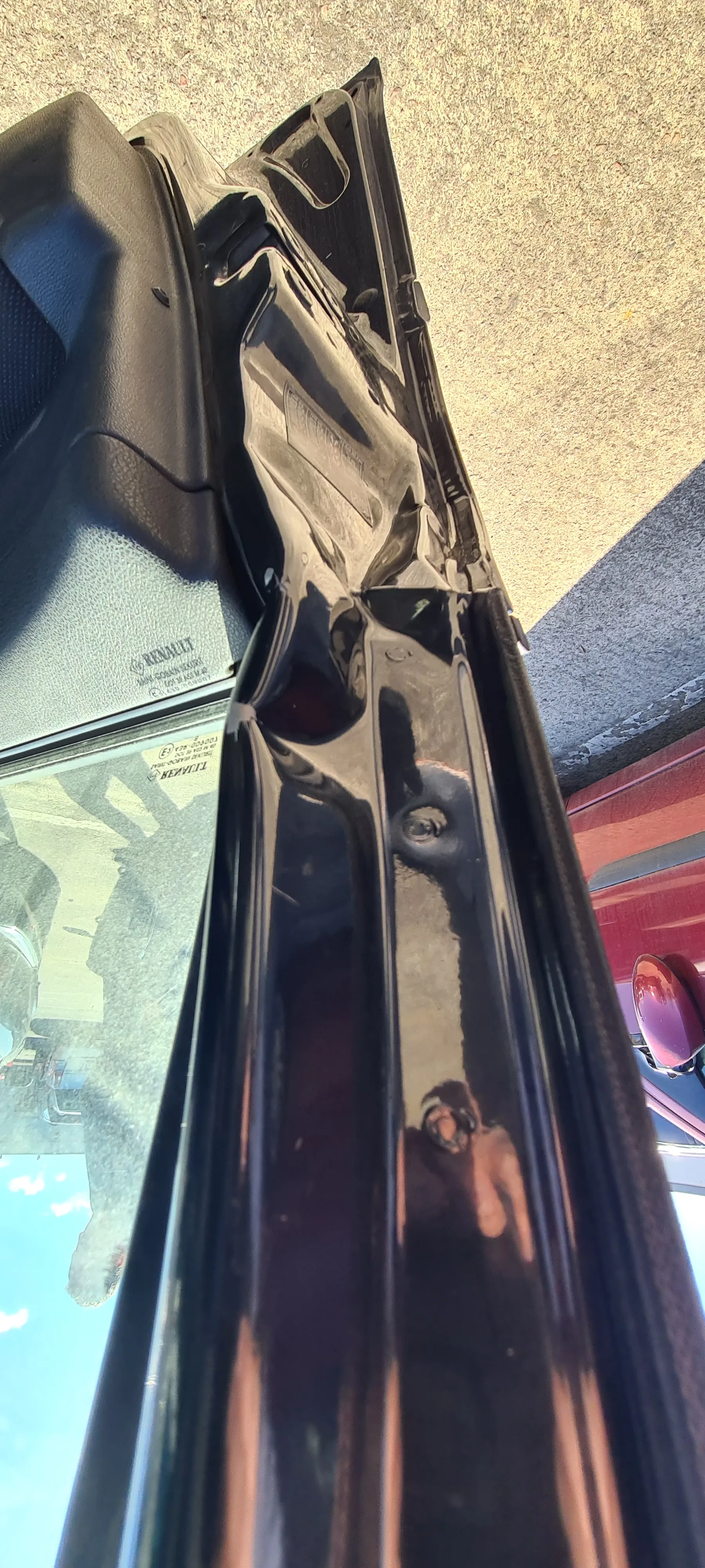 Zbliżenie na czarną, połyskującą krawędź drzwi samochodu z częścią szyby. Na szybie widoczny jest odbicie budynków, nieba oraz osoby fotografującej. W tle fragment czerwonego samochodu i asfaltowej nawierzchni. Na szybie widoczny napis 'TUV' oraz inne nieczytelne znaki.