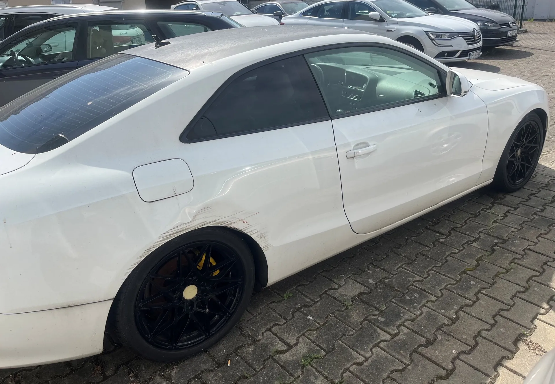 Biały samochód osobowy typu coupe zaparkowany na kostce brukowej. Pojazd posiada widoczne uszkodzenia karoserii w postaci wgnieceń i zarysowań nad tylnym lewym kołem. Samochód ma czarne felgi z żółtymi zaciskami hamulcowymi. W tle widoczne są inne zaparkowane samochody w kolorach białym i szarym.