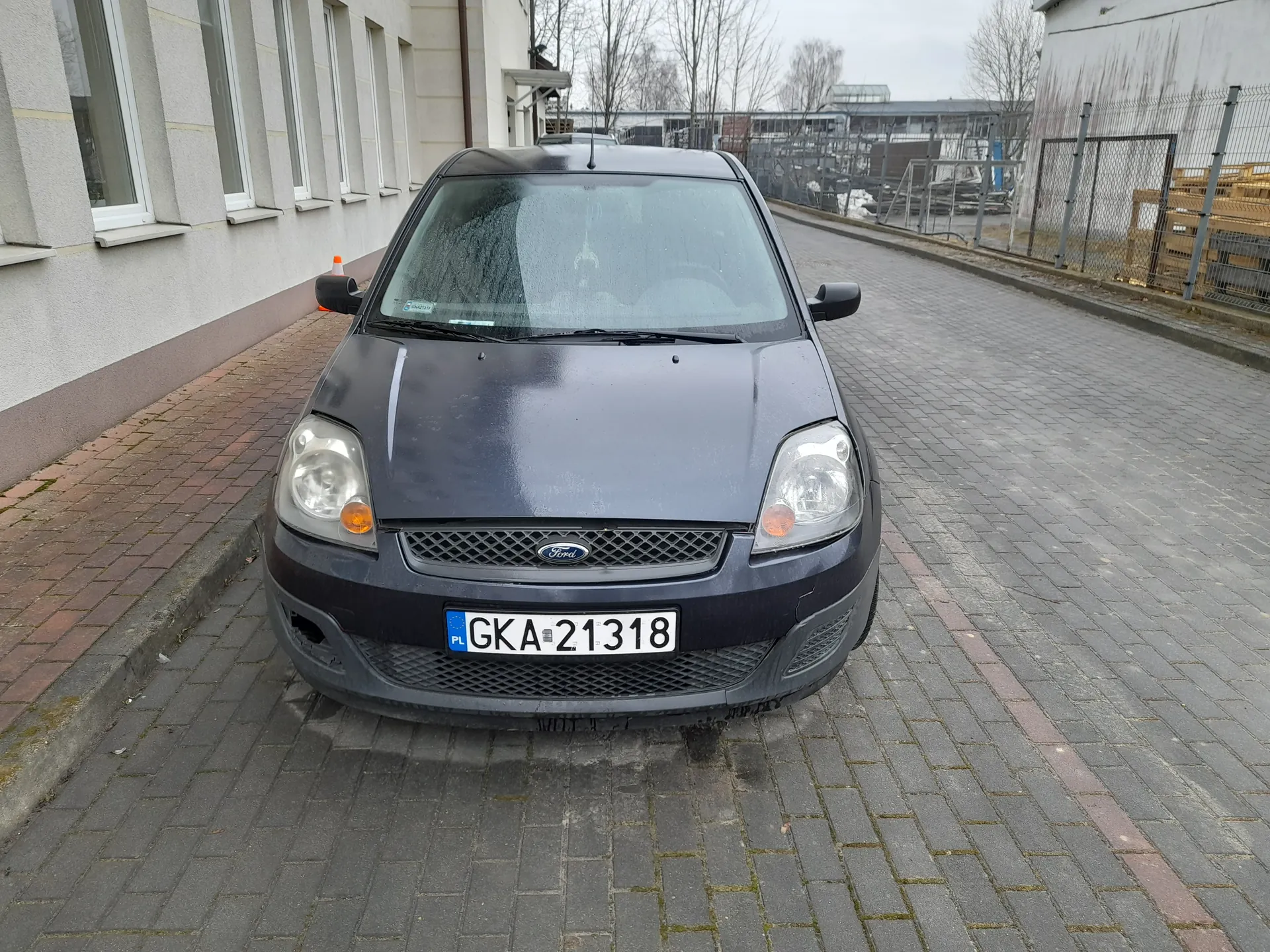Ciemnoszary samochód marki Ford zaparkowany na brukowanej drodze. Widoczna tablica rejestracyjna PL GKA-21318. Po lewej stronie budynek z oknami, po prawej ogrodzenie i przemysłowe konstrukcje. Tło z drzewami bez liści i pochmurnym niebem.