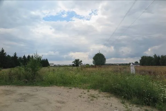 Krajobraz z piaszczystą drogą na pierwszym planie, przechodzącą w dziką roślinność i trawę. W oddali widoczne są drzewa, kilka niskich budynków oraz słupy energetyczne z liniami napowietrznymi. Niebo jest w większości zachmurzone z niewielką przerwą ukazującą błękit.