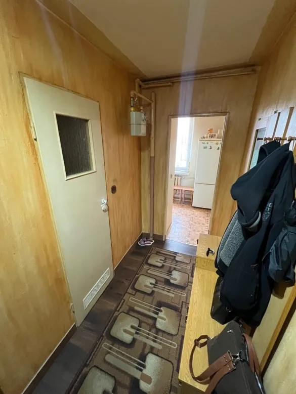 Wnętrze pomieszczenia z drewnianymi ścianami i podłogą. W centrum znajduje się jasnobrązowe drzwi z małym prostokątnym okienkiem. Nad drzwiami umieszczono lustrzane lustro odbijające fragment kuchni z białymi szafkami i oknem. Po prawej stronie drzwi leży dywan z geometrycznym wzorem w brązowych odcieniach. Na drewnianym stole po prawej stronie widoczna jest ciemna kurtka oraz brązowa torebka. W pobliżu lustra stoją małe przedmioty, w tym fioletowy obiekt i białe pudełko. Ściana po lewej stronie jest jednolita, bez dodatkowych elementów.