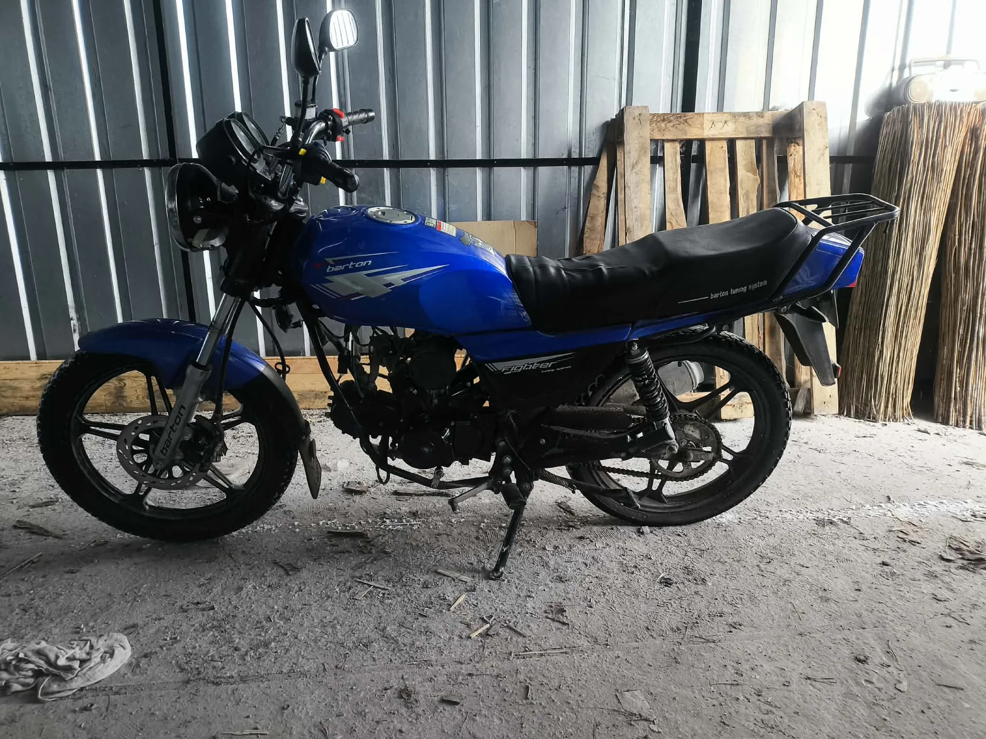 Niebieski motocykl marki Barton z napisem 'fighter' na boku, czarnym siedzeniem z napisem 'barton tuning system' oraz widocznym napisem 'barton' na przednim widelcu. Motocykl stoi na podpórce w pomieszczeniu z metalowymi ścianami, betonową podłogą z rozsypanymi drobnymi przedmiotami oraz drewnianymi paletami i stosem suchych łodyg w tle.