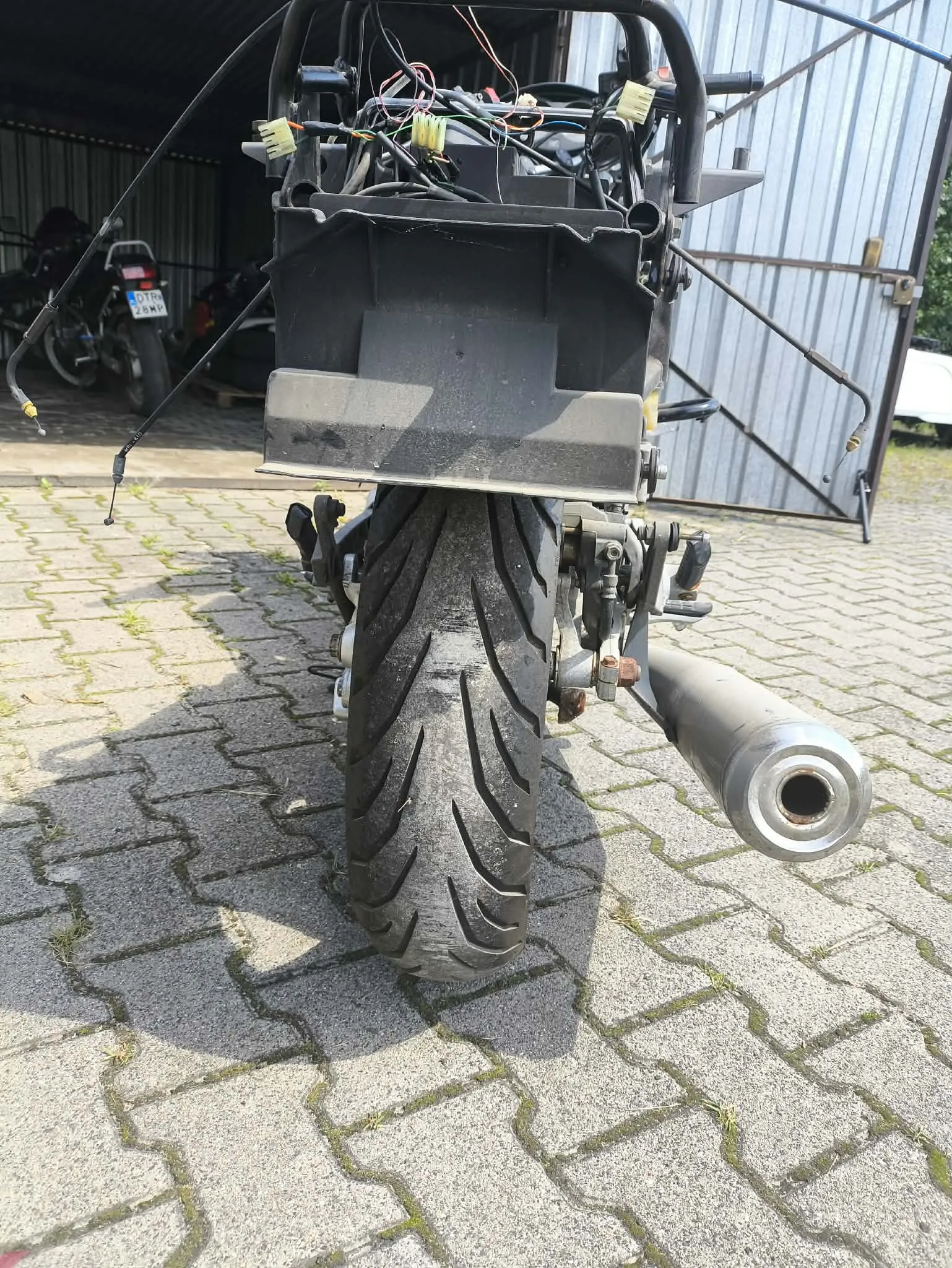 Widok z tyłu na motocykl zaparkowany na kostce brukowej. Widoczna jest tylna opona z wyraźnym bieżnikiem, rura wydechowa po prawej stronie oraz tylny błotnik i stelaż z widocznymi przewodami elektrycznymi. W tle znajduje się metalowa konstrukcja garażowa oraz fragment innego motocykla z tablicą rejestracyjną.