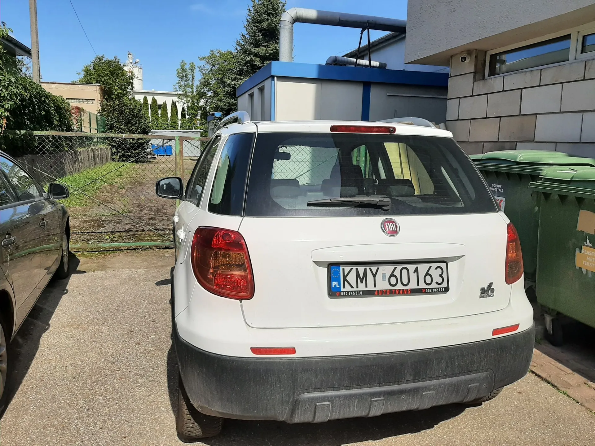 Tylny widok białego samochodu marki Fiat 500L zaparkowanego na podjeździe. Pojazd ma polską tablicę rejestracyjną o numerze KMY 60163. W tle widoczny jest niebieski budynek techniczny z rurami na dachu, zielone kontenery na odpady oraz fragment innego, srebrnego samochodu po lewej stronie.