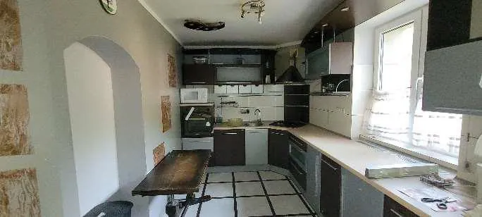 Kuchnia z białymi i ciemnobrązowymi szafkami, drewnianym stołem w centrum, podłogą z białymi płytkami i czarnymi liniami. Po prawej stronie duże okno z widokiem na zewnątrz, na ścianie po lewej łukowate wnęka i fragmenty starannie usuniętej tapety. W tle widoczne urządzenia: mikrofalówka, zmywarka, kuchenka gazowa oraz czarna kloszowa kuchenna. Sufit z wiszącym oświetleniem i okrągłym zegarem na ścianie.