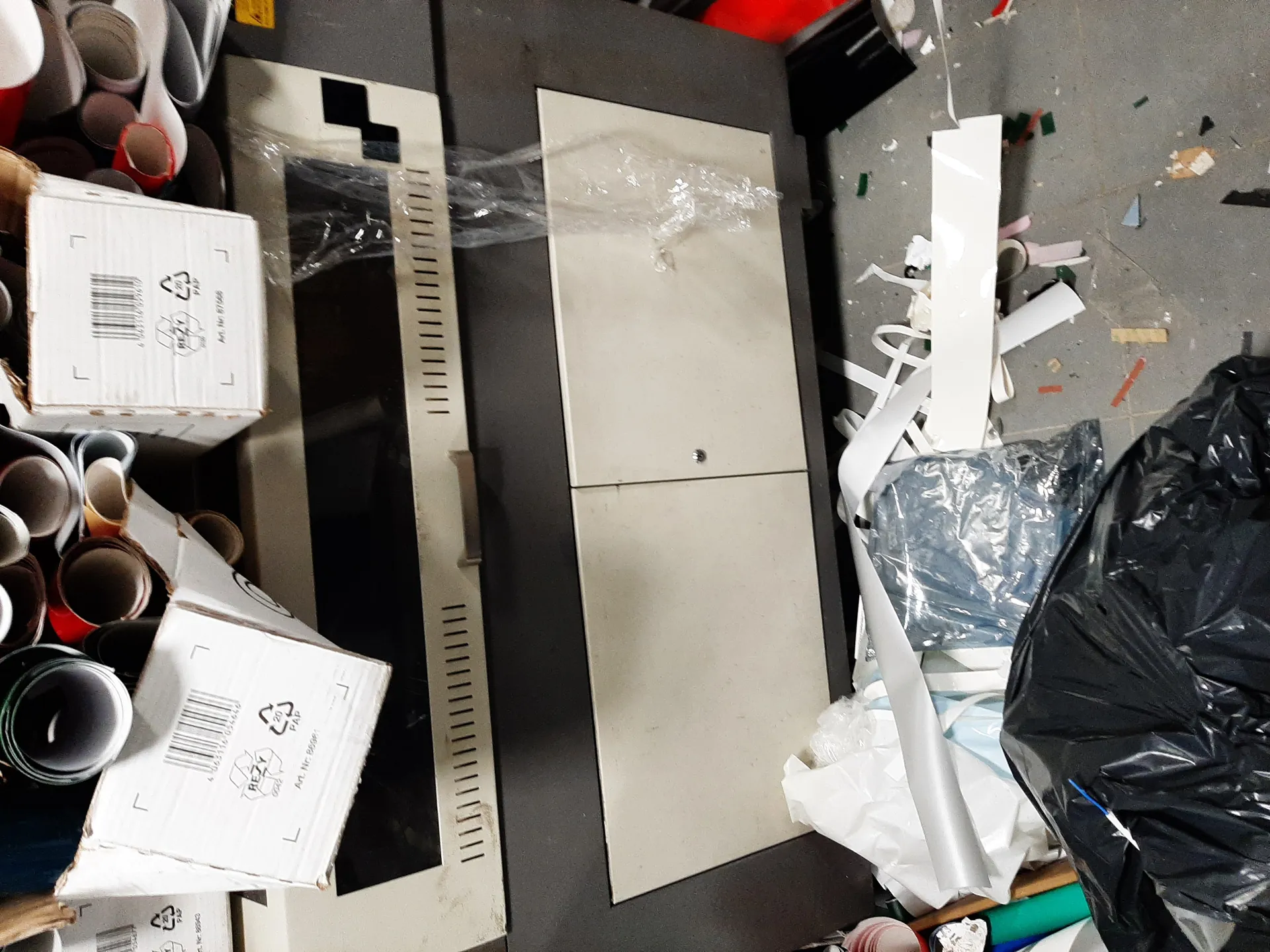 Zdjęcie przedstawia przestrzeń roboczą z różnymi przedmiotami. Po lewej stronie znajdują się kartonowe pudełka z kodami kreskowymi, symbolami recyklingu oraz widocznym tekstem 'REZY'. Obok nich leżą zwinięte rulony materiału w kolorach szarym i czerwonym. W centrum obrazu umieszczono ciemnoszary szafę lub urządzenie z jasnymi panelami, częściowo przykryte przezroczystą folią. Po prawej stronie widoczne są białe paski materiału, czarna torba na śmieci oraz rozrzucone na podłodze drobne elementy i śmieci. Podłoga jest szara, z widocznymi plamami i kawałkami materiału.