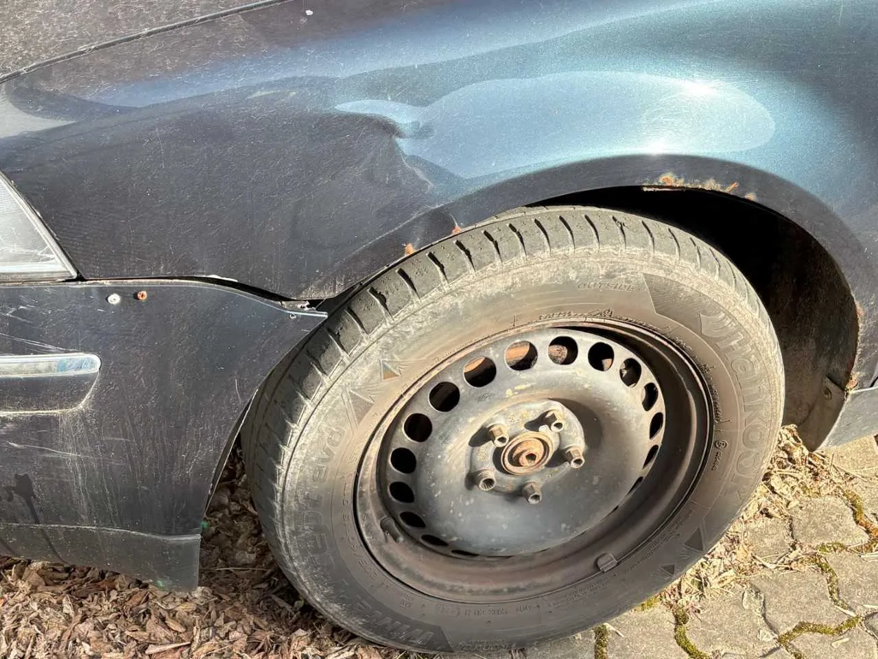 Zbliżenie na przód ciemnego samochodu z uszkodzonym lewym przednim błotnikiem. Widoczna zdeformowana karoseria, zarys reflektora i kierunkowskazu. Opona z brudnym, metalowym felgiem o otworach w kształcie okręgów, z widocznym rdzą na śrubach i osi. Podwozie oparte na podłożu z suchych liści i brukowanych płyt z mchem. Opona ma czytelne napisy, w tym 'CITY' i '205/55 R16'.