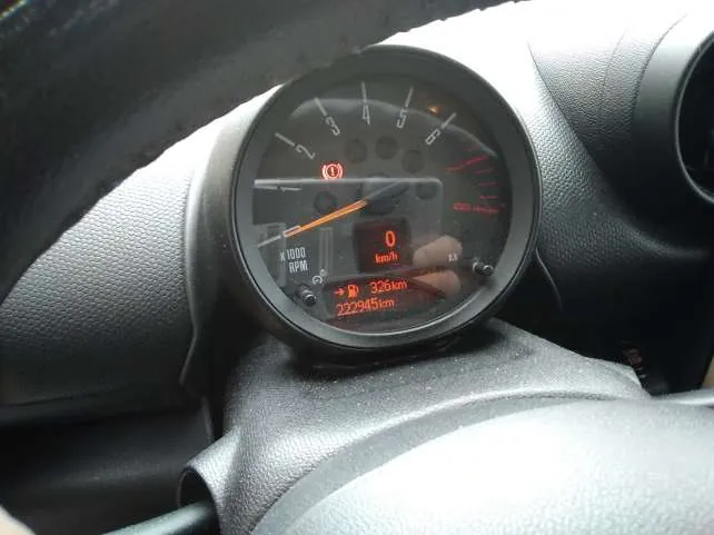 Zbliżenie na zegar samochodowy z czarnym tłem. Wskazówka prędkościomierza wskazuje 0 km/h, na wyświetlaczu cyfrowym widoczne są wartości: 326 km oraz 222945 km. Obecny jest czerwony sygnalizator ostrzegawczy z wykrzyknikiem. W tle fragment kierownicy i deski rozdzielczej z czarną, teksturowaną powierzchnią.