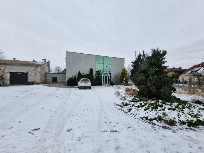 Zimowy krajobraz z pokrytą śniegiem drogą i terenem. W centrum widoczny nowoczesny budynek o szarych ścianach i zielonym szklanym elewacją. Przed budynkiem stoi biały samochód typu SUV. Po lewej stronie starszy, kamienny budynek z bramą garażową. Wokół rosną drzewa i krzewy, częściowo pokryte śniegiem. Niebo jest zachmurzone. W tle widoczne są dodatkowe domy i linie energetyczne.