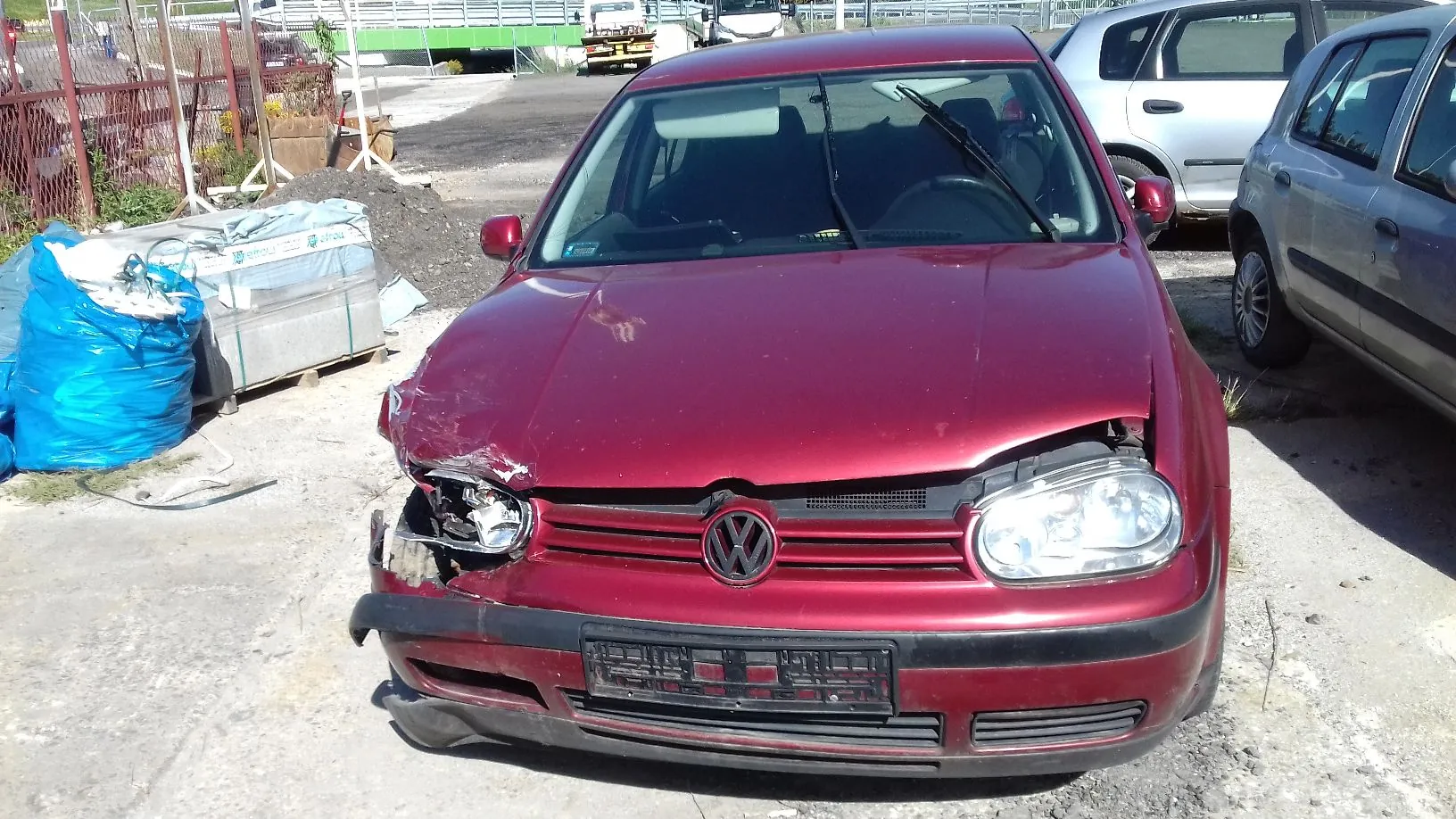 Zdjęcie przodu uszkodzonego, bordowego samochodu marki Volkswagen. Pojazd ma wgniecioną maskę po prawej stronie (z perspektywy widza po lewej) oraz rozbite przednie światło z tej samej strony. Samochód stoi na betonowym placu obok niebieskiej plandeki i innych pojazdów.