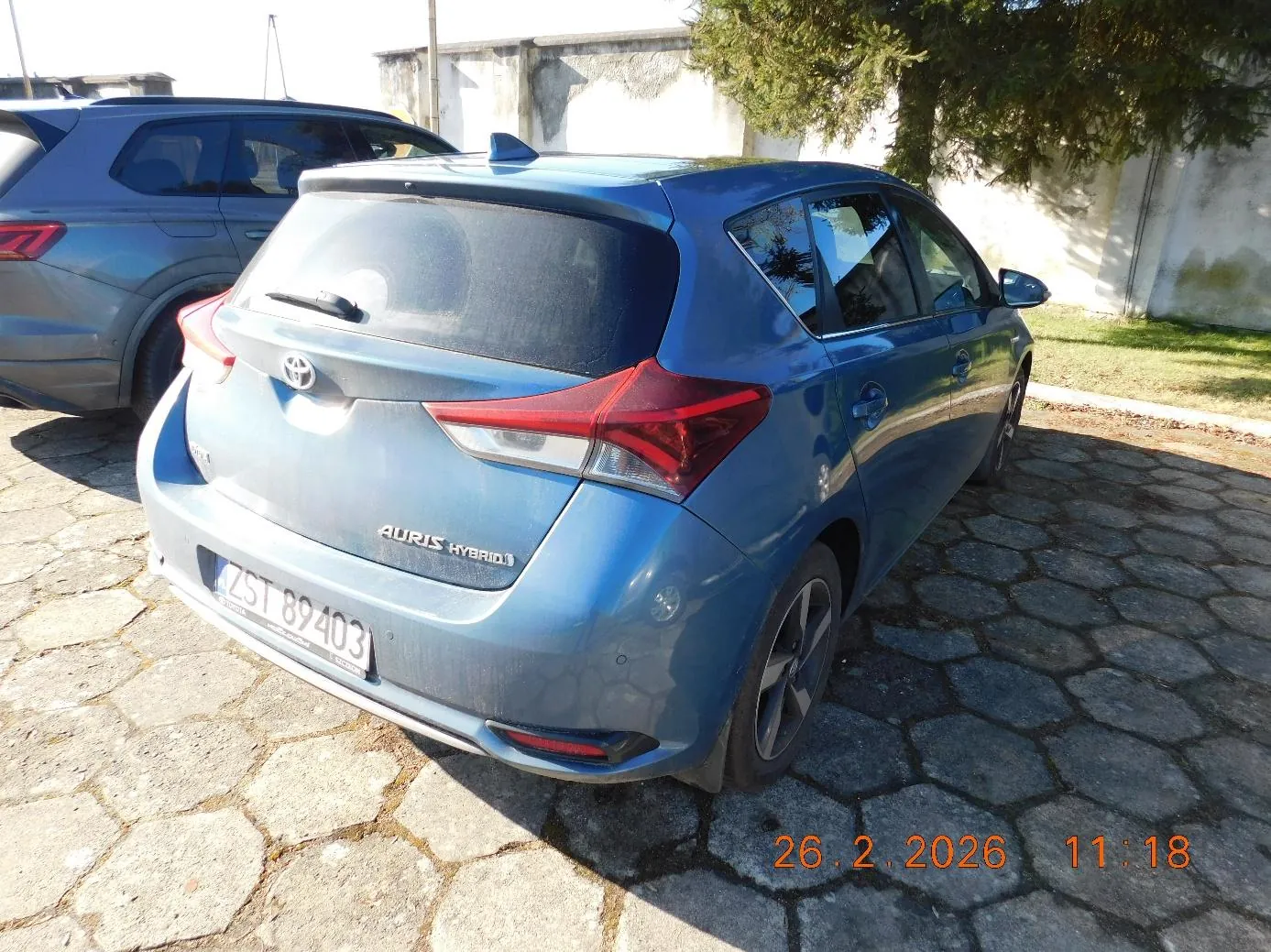Niebieski samochód Toyota Auris Hybrid widziany od tyłu, z tablicą rejestracyjną ZST 89403, zaparkowany na brukowanej powierzchni. W tle szary SUV, biała ściana, drzewa i trawa. Na obrazie widoczny znaczek daty i godziny: 26.2.2026 11:18.