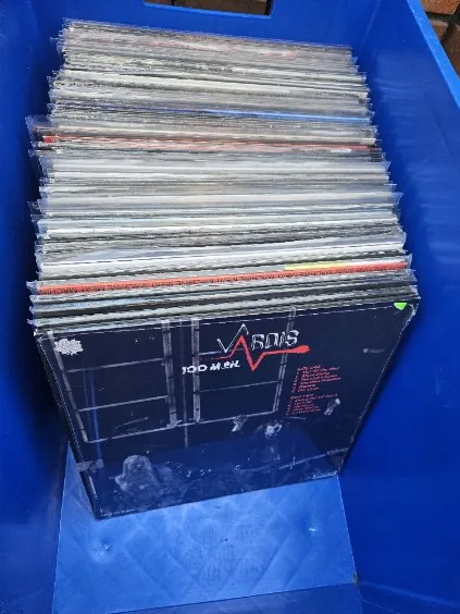 Stos winylowych płyt w niebieskim plastikowym pojemniku. Na górze stosu widoczna okładka z napisem "VOL 100 M 84" oraz czerwonym logo. Okładka ma ciemne tło z siatką i małym obrazkiem postaci.