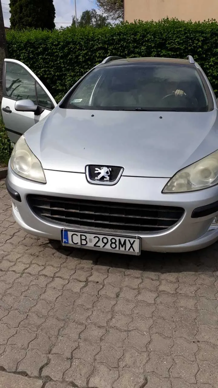 Srebrny samochód marki Peugeot z widocznym logo na masce, z tablicą rejestracyjną CB 298MX z polskim oznaczeniem PL po lewej stronie. Drzwi pasażera po lewej stronie są otwarte, a wewnątrz widoczna jest osoba trzymająca kierownicę. Tło stanowi zielony żywopłot, drzewa i fragment budynku. Samochód stoi na brukowanej powierzchni.