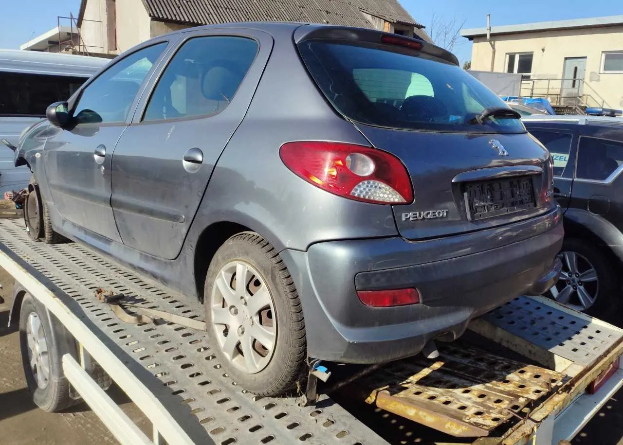 Szary samochód Peugeot na metalowej platformie holownika. Widok od tyłu z widocznym logo i napisem 'PEUGEOT' na bagażniku. Samochód ma zabrudzoną karoserię, a na platformie widoczne są otwory i ślady korozji. W tle inne pojazdy, budynki oraz niebieskie niebo.