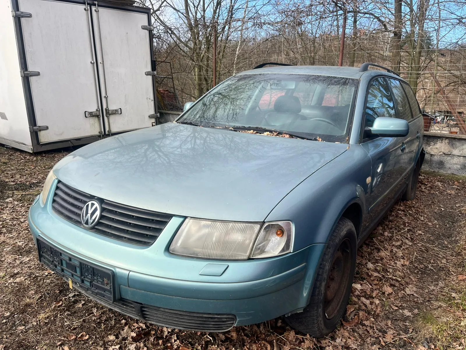 Niebieski samochód osobowy marki Volkswagen zaparkowany na podwórku pokrytym suchymi liśćmi. Na masce widoczne są liście, a miejsce na tablicę rejestracyjną jest puste. W tle biała przyczepa z zamkniętymi drzwiami, drzewa bez liści oraz ogrodzenie. Samochód ma zarysowaną linię dachu i zatłuszczoną przednią szybę.