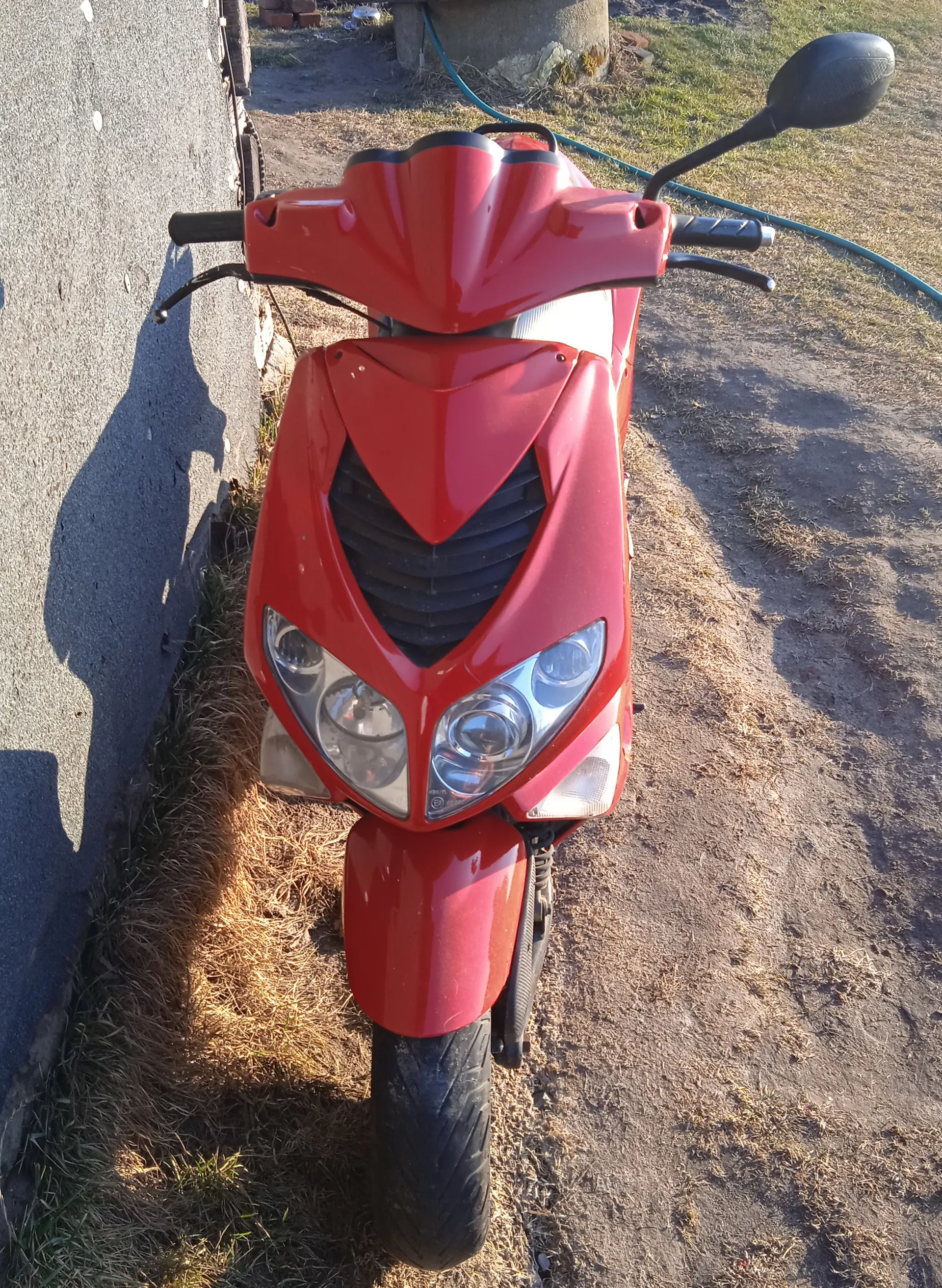 Czerwony skuter widziany od przodu z czarnymi akcentami, dwoma reflektorami, czarnymi kierownicami z lusterkami. Po lewej stronie betonowa ściana, na tle sucha trawa i ziemia, niebieski wąż.