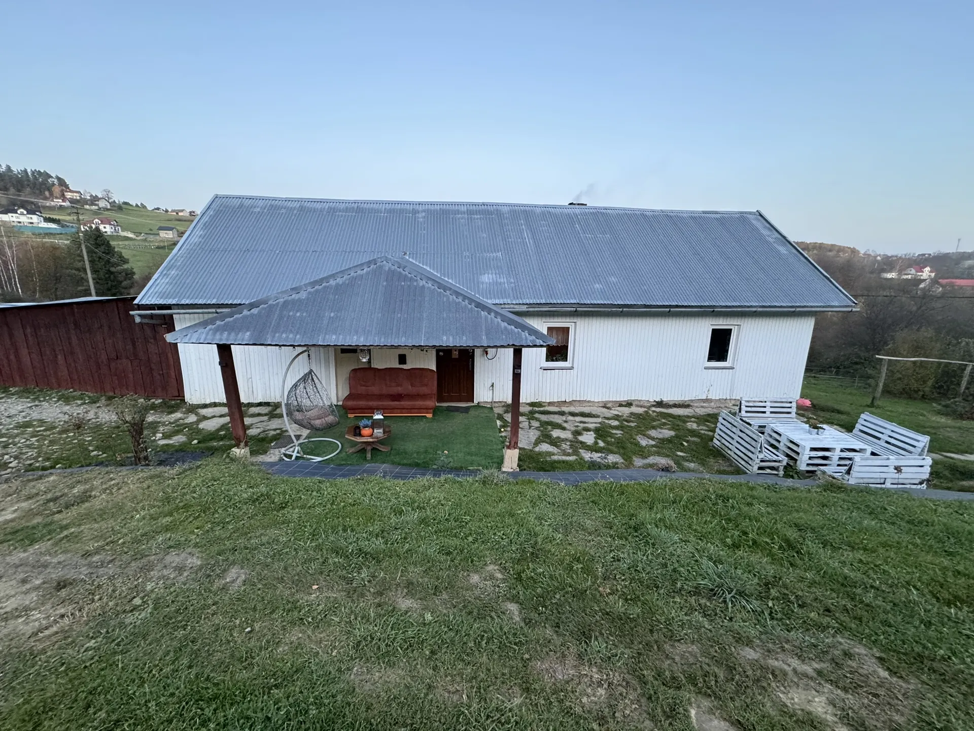 Biały dom z metalowym dachem w kolorze szarym. Przed domem znajduje się daszek z brązową sofą, wiszącym fotelikiem i małym stolikiem z przedmiotami. Po prawej stronie domu leżą białe palety drewniane. Wokół domu trawa z miejscami odsłoniętej ziemi. Po lewej stronie widoczny brązowy płot i drzewa. W tle domy, wzgórza i jasne niebo bez chmur.