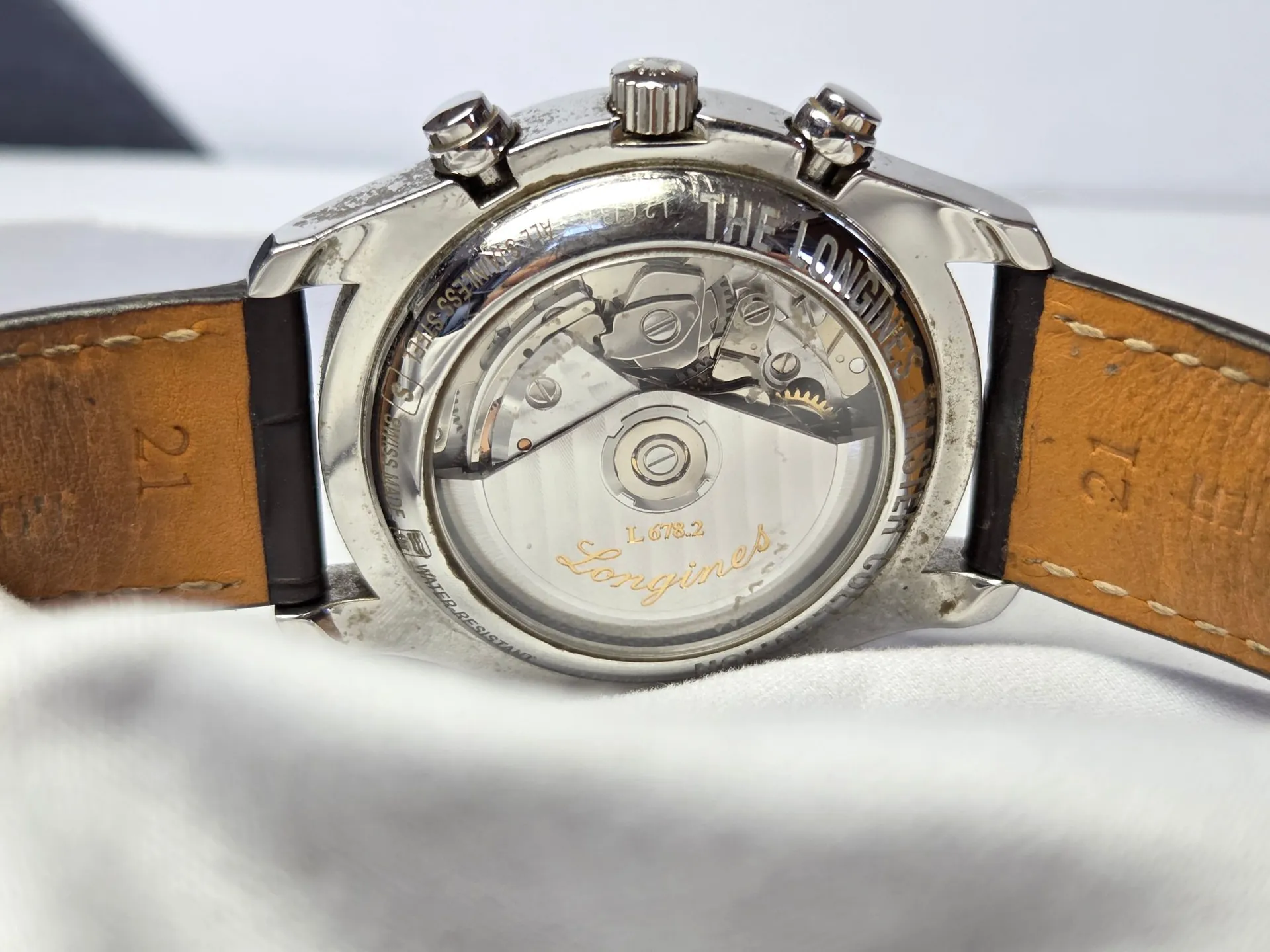 Zdjęcie tyłu srebrnego zegarka marki Longines z przezroczystym deklem, przez który widoczny jest mechanizm. Na szklanym dnie znajduje się napis 'L678.2' oraz charakterystyczne logo 'Longines'. Zegarek jest przymocowany do brązowego, skórzanego paska. Widoczne są trzy pokrętła na górze koperty oraz grawerunek na obwodzie tarczy: 'WATER RESISTANT' i 'SWISS MADE'.
