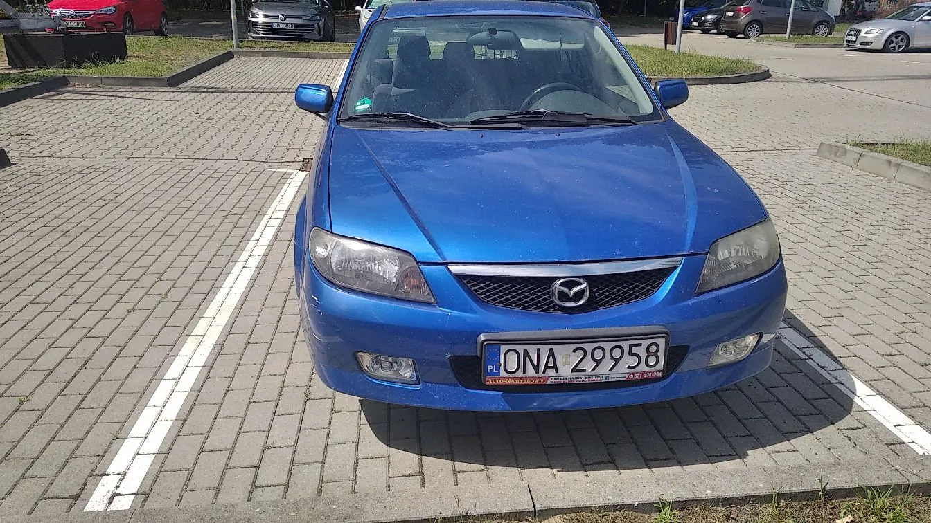 Niebieski samochód osobowy marki Mazda zaparkowany na kostce brukowej. Widoczny jest przód pojazdu z tablicą rejestracyjną o numerze ONA 29958. W tle znajdują się inne zaparkowane samochody oraz krawężniki wyznaczające miejsca parkingowe.