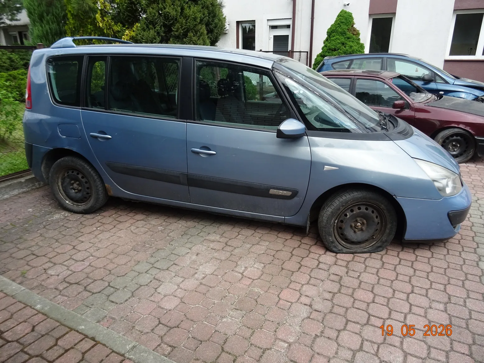 Niebieski samochód typu minivan zaparkowany bokiem na kostce brukowej. W tle widoczne są inne zaparkowane samochody w kolorach bordowym i niebieskim oraz fragment białego budynku z oknami i zielonymi krzewami. W prawym dolnym rogu zdjęcia znajduje się czerwona data: 19 05 2026.