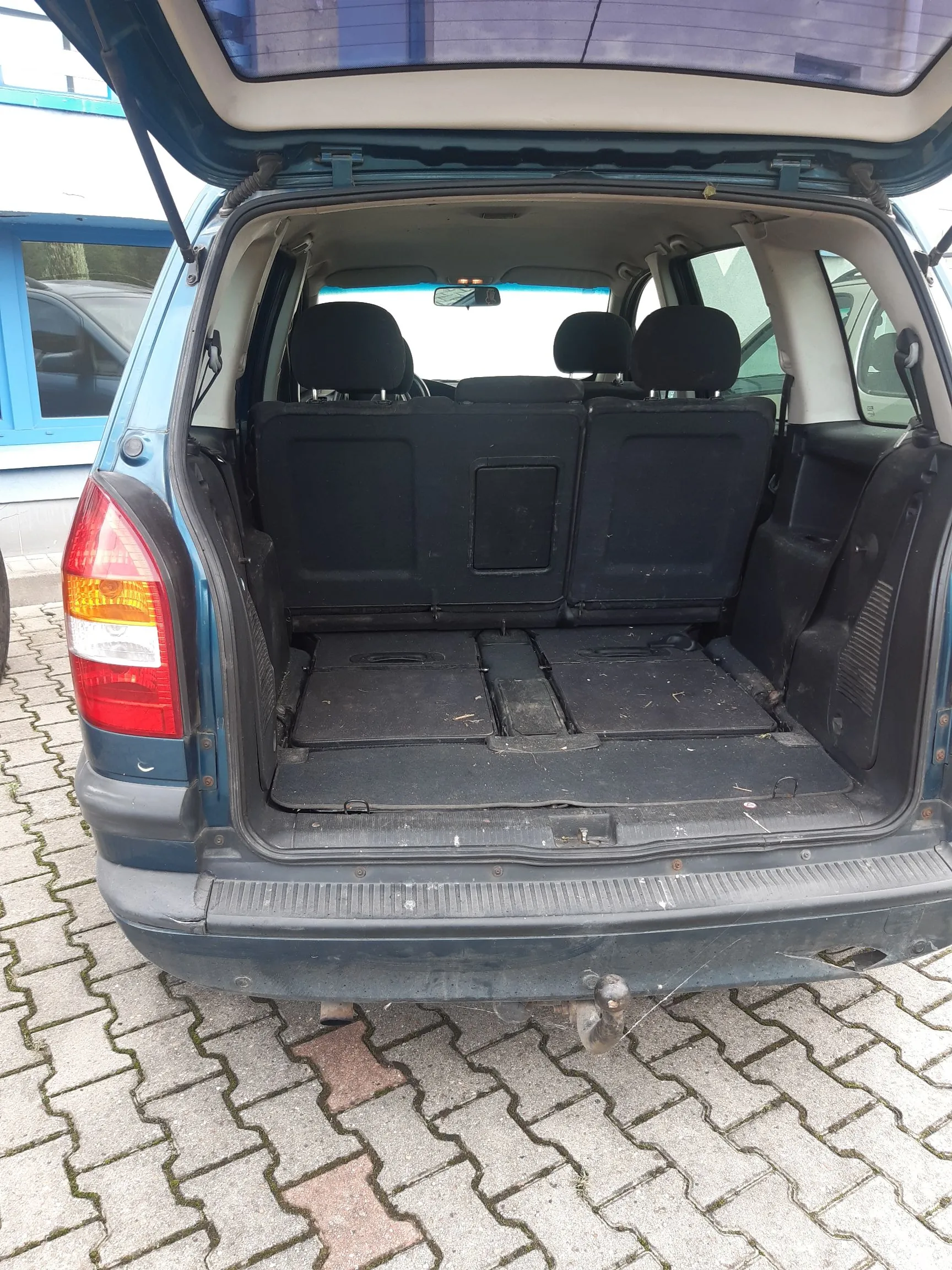 Widok z tyłu do otwartego bagażnika niebieskiego samochodu. Wnętrze bagażnika jest wyłożone ciemnoszarym materiałem, a tylne siedzenia są złożone, tworząc płaską powierzchnię. W tle widoczne są zagłówki przednich i tylnych foteli. Samochód zaparkowany jest na kostce brukowej.