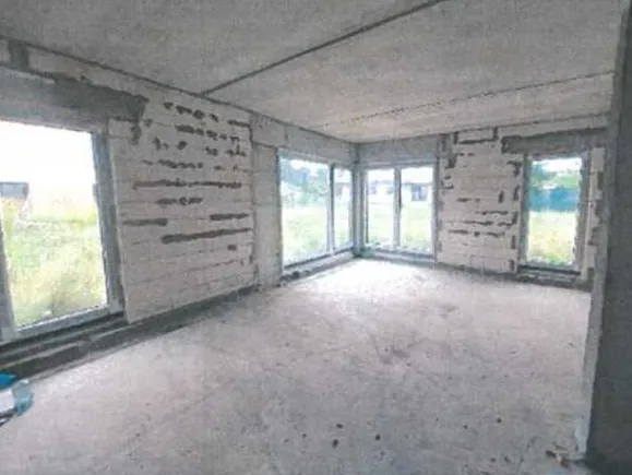 Pusty pomieszczenie z betonowymi ścianami i podłogą. Wiele dużych okien po bokach pomieszczenia, przez które widać zieloną trawę i budynki na zewnątrz. Ściany mają odsłonięte fragmenty, gdzie brakuje tynku. Na podłodze w lewym dolnym rogu znajduje się niebieski przedmiot. Sufit jest betonowy, a całość pomieszczenia wygląda na niedokończoną lub nieużywaną.