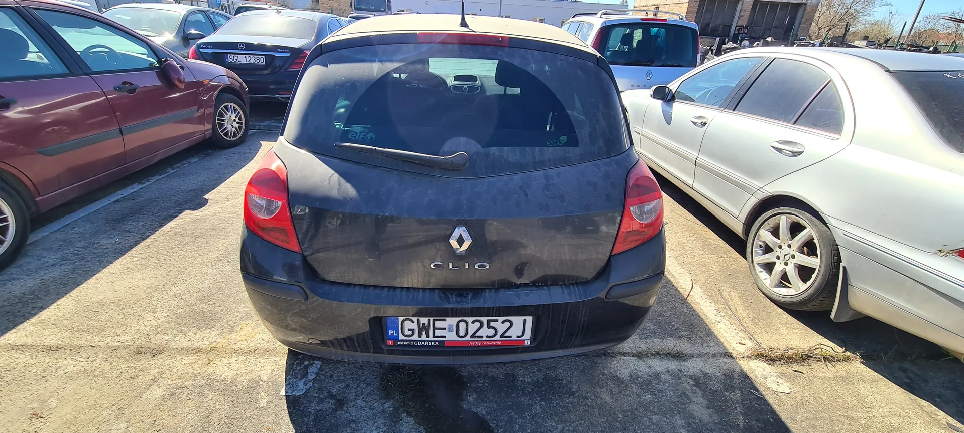 Zdjęcie przedstawia czarny samochód Renault Clio zaparkowany na placu parkingowym. Widok z tyłu, z widoczną tablicą rejestracyjną PL GWE 0252 J oraz napisem CLIO pod logo marki. Po obu stronach znajdują się inne pojazdy: czerwony samochód po lewej i srebrny Mercedes po prawej. W tle widoczne są budynki, drzewa oraz inne zaparkowane samochody. Na szybie tylnej Clio znajdują się naklejki z napisami elf i eco. Asfaltowy plac parkingowy z białymi liniami wyznaczającymi miejsca postojowe.