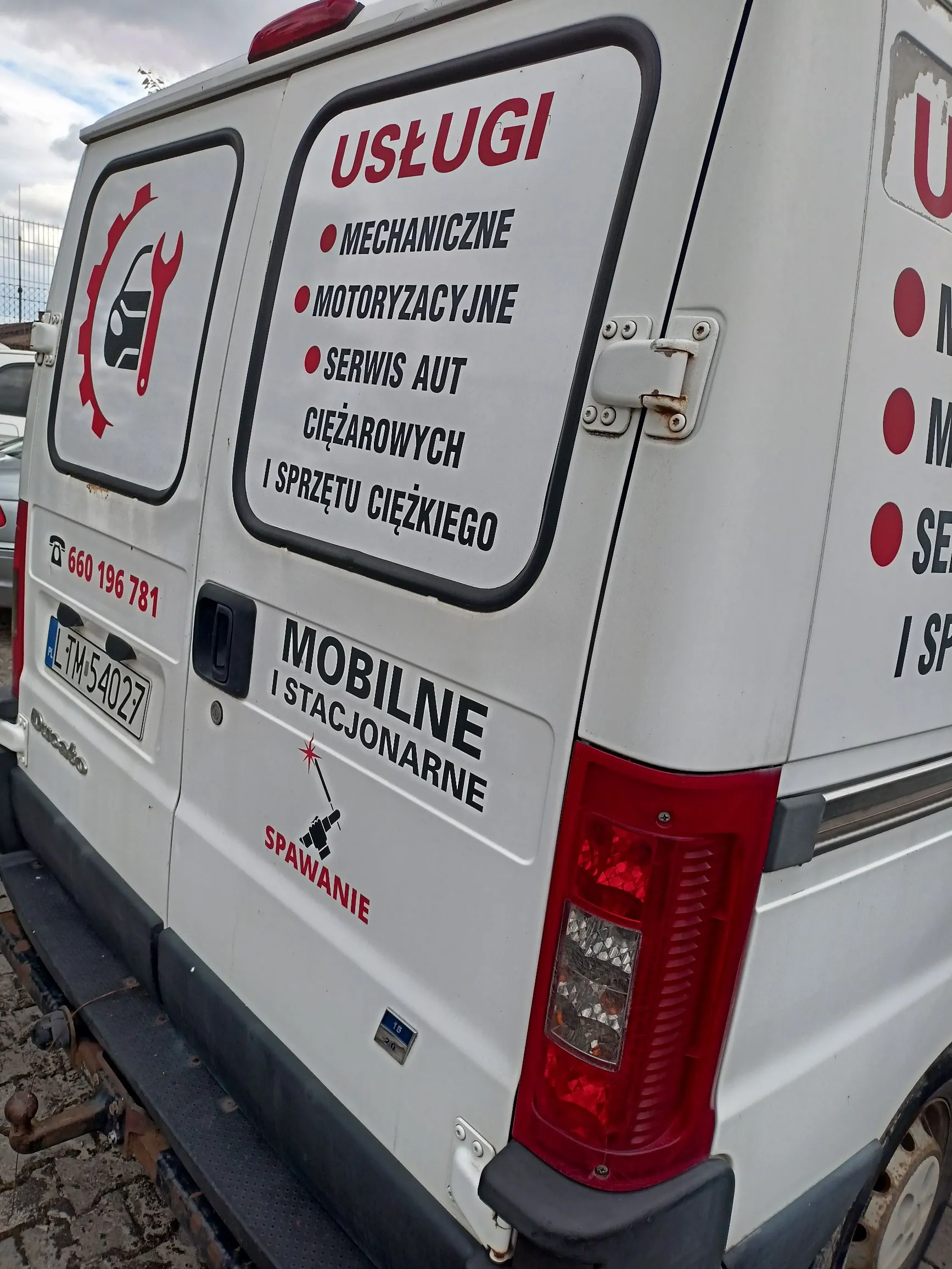 Tylna część białego samochodu dostawczego z reklamami usług. Na tylnych drzwiach znajdują się napisy: 'USŁUGI', 'MECHANICZNE', 'MOTORYZACYJNE', 'SERWIS AUT CIĘŻAROWYCH I SPRZĘTU CIĘŻKIEGO'. Poniżej widnieje napis 'MOBILNE I STACJONARNE' oraz 'SPOWANIE' obok grafiki spawarki. Po lewej stronie znajduje się numer telefonu '660 196 781' oraz tablica rejestracyjna 'LTM 54027'. Po lewej stronie widoczne jest logo z sylwetką samochodu i kluczem płaskim w czerwonym kole.