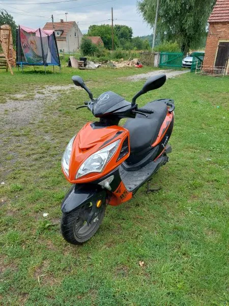Pomarańczowo-czarny skuter zaparkowany na trawiastym terenie. W tle widoczny jest dom, zielone ogrodzenie oraz fragmenty roślinności.