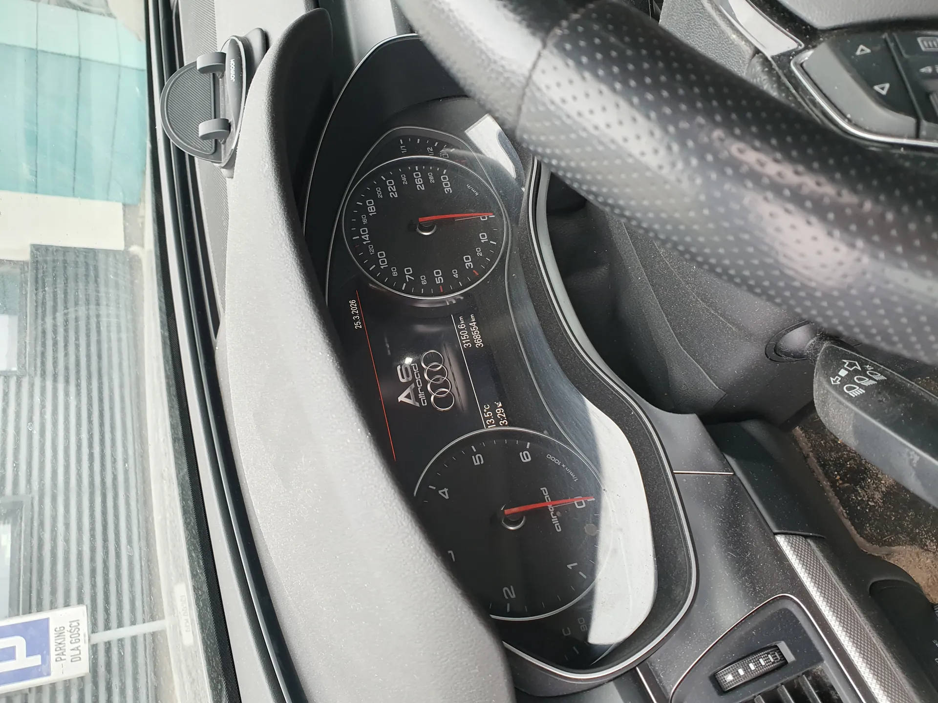 Wnętrze samochodu Audi A6 z widoczną deską rozdzielczą. Licznik prędkości z zakresem do 300 km/h, obrotomierz, wyświetlacz cyfrowy z napisem „A6” i logo Audi. Przebieg 3150,6 km, temperatura zewnętrzna 13,5°C, data 25.3.2026. Bieżący bieg: P (parking). Po lewej stronie przez okno widoczny znak „PARKING DLA GOSCI” oraz fragment budynku z szklaną elewacją. Kierownica po prawej stronie.