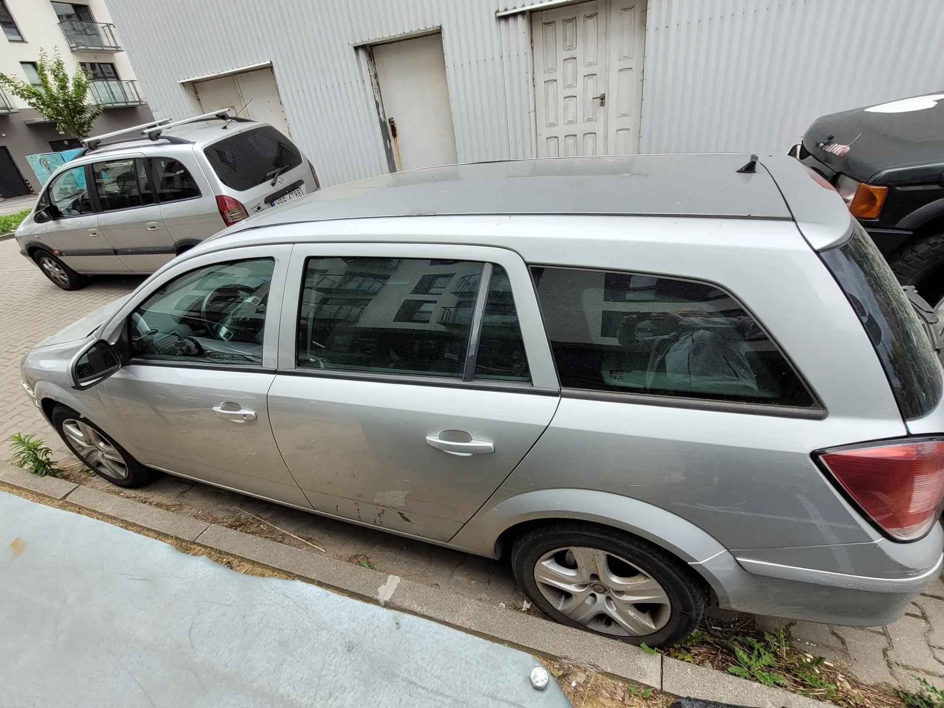 Srebrny samochód typu kombi zaparkowany przy chodniku na brukowanej ulicy. W tle srebrny minivan i czarny SUV. Budynek z białą, pionowo listwowaną elewacją, białymi drzwiami i małymi oknami. Na chodniku widoczne są fragmenty trawy i betonowa krawężnik. Samochody stoją na płaskim terenie, horyzont jest poziomy.