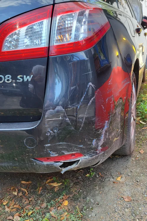 Zbliżenie na tylny prawy róg ciemnego samochodu, który posiada widoczne uszkodzenia. Na zderzaku i błotniku znajdują się liczne zarysowania, otarcia oraz wgniecenia. W miejscu uszkodzeń odsłonięta jest czerwona warstwa podkładu lub element konstrukcyjny. Dolna część zderzaka jest pęknięta i częściowo oderwana. Widoczny jest fragment tylnej lampy oraz fragment napisu '08.SW' na klapie bagażnika.