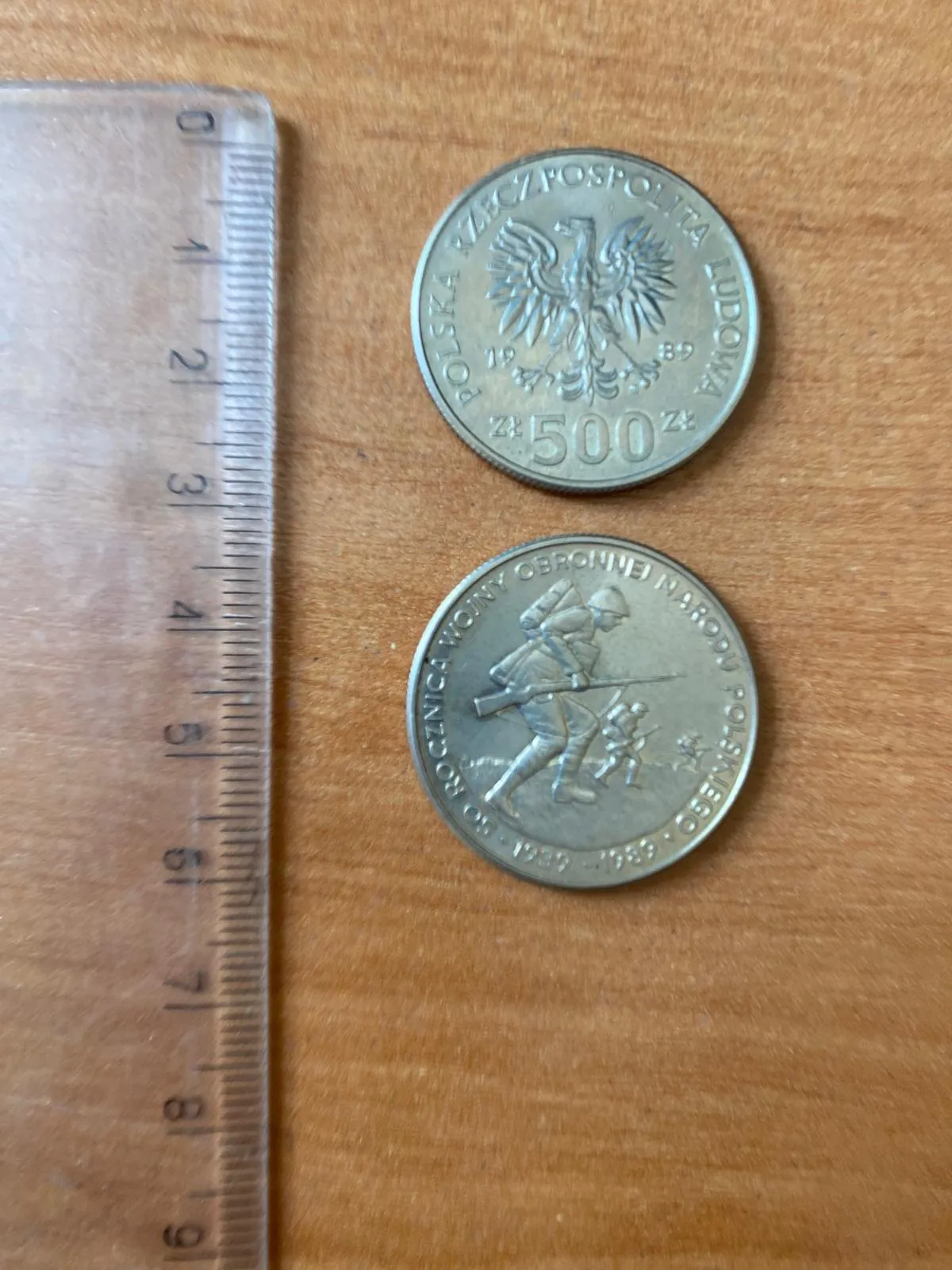 Zdjęcie przedstawiające dwie strony polskiej monety o nominale 500 zł z roku 1989, leżące na drewnianym blacie obok plastikowej linijki. Górna moneta pokazuje awers z godłem Polski (orłem) i napisem „POLSKA RZECZPOSPOLITA LUDOWA” oraz nominałem „500 zł”. Dolna moneta pokazuje rewers z postacią żołnierza w ruchu z karabinem i napisem „ROcznica WOJNY OBRONNEJ NARODU POLSKIEGO 1939-1989”. Linijka po lewej stronie wskazuje rozmiar monety, który wynosi około 2,5 cm w średnicy.