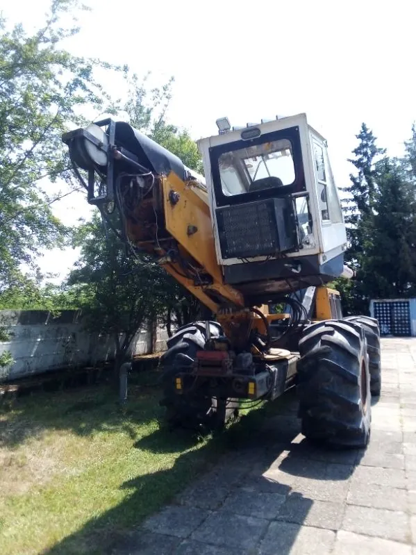 Żółto-biała koparka z dużymi, traktorowymi oponami, zaparkowana na betonowej ścieżce obok trawnika i drzew. Maszyna posiada białą kabinę operatora oraz wysięgnik w kolorze żółtym.