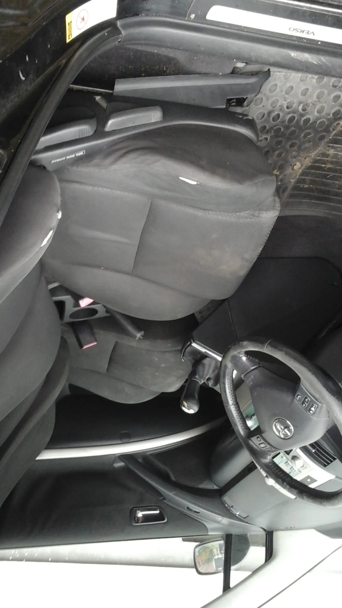 Wnętrze samochodu z czarnymi siedzeniami, kierownicą z logo Toyoty, dźwignią zmiany biegów i drzwiami kierowcy. Widoczne są uchwyt na drzwiach, lusterko boczne oraz napis "VERSO" na ramie drzwi. Podłoga pokryta jest czarną mata z otworami, a na siedzeniach i materiale widoczne są ślady brudu. W tle fragment przedniej szyby i deski rozdzielczej.