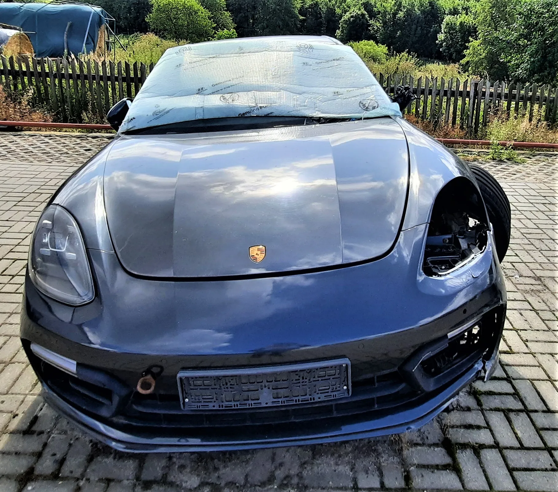 Przód ciemnoniebieskiego samochodu marki Porsche z widocznym logo na masce. Pojazd jest uszkodzony: brakuje lewego reflektora, a przedni zderzak jest częściowo zniszczony. Przednia szyba jest zaklejona jasnoniebieską folią ochronną. Samochód stoi na kostce brukowej przed drewnianym płotem i zielonymi drzewami.