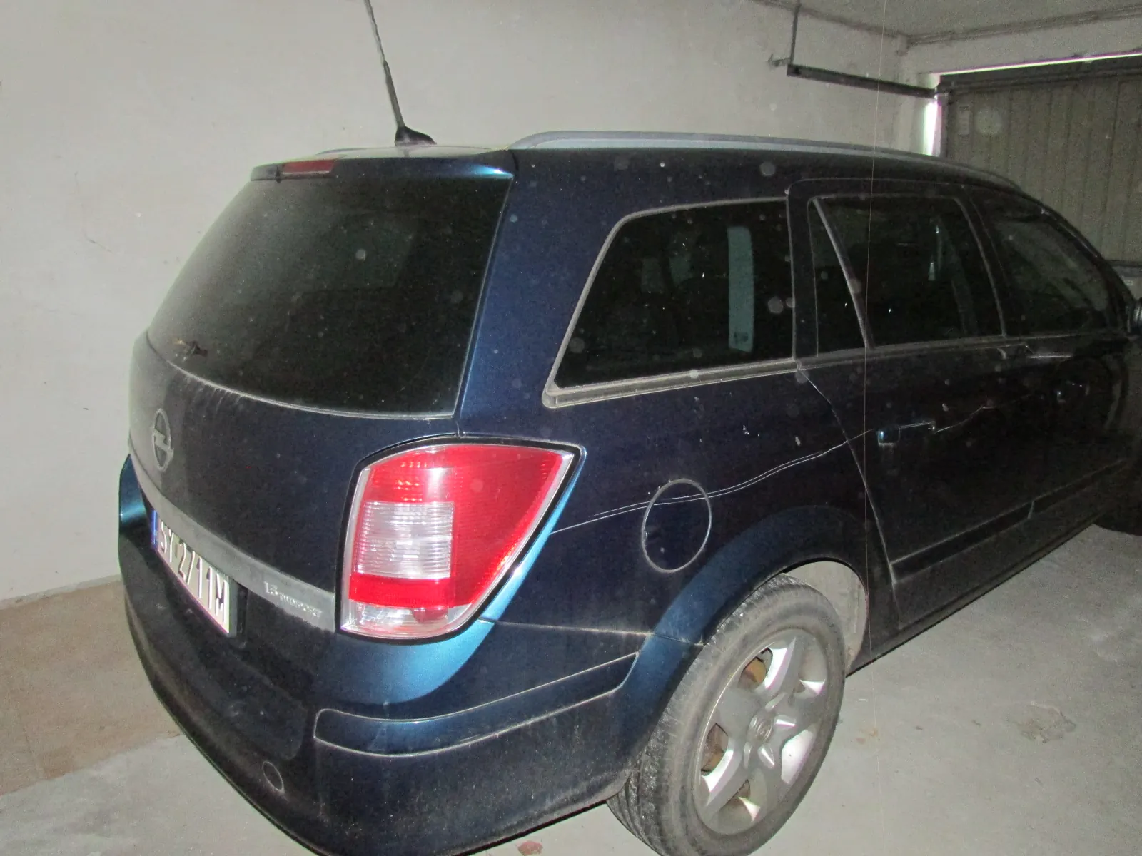 Tylna i boczna część ciemnoniebieskiego samochodu typu kombi marki Opel, zaparkowanego w jasnym pomieszczeniu. Samochód jest zakurzony, widoczna jest tylna lampa, antena na dachu oraz fragment tablicy rejestracyjnej.