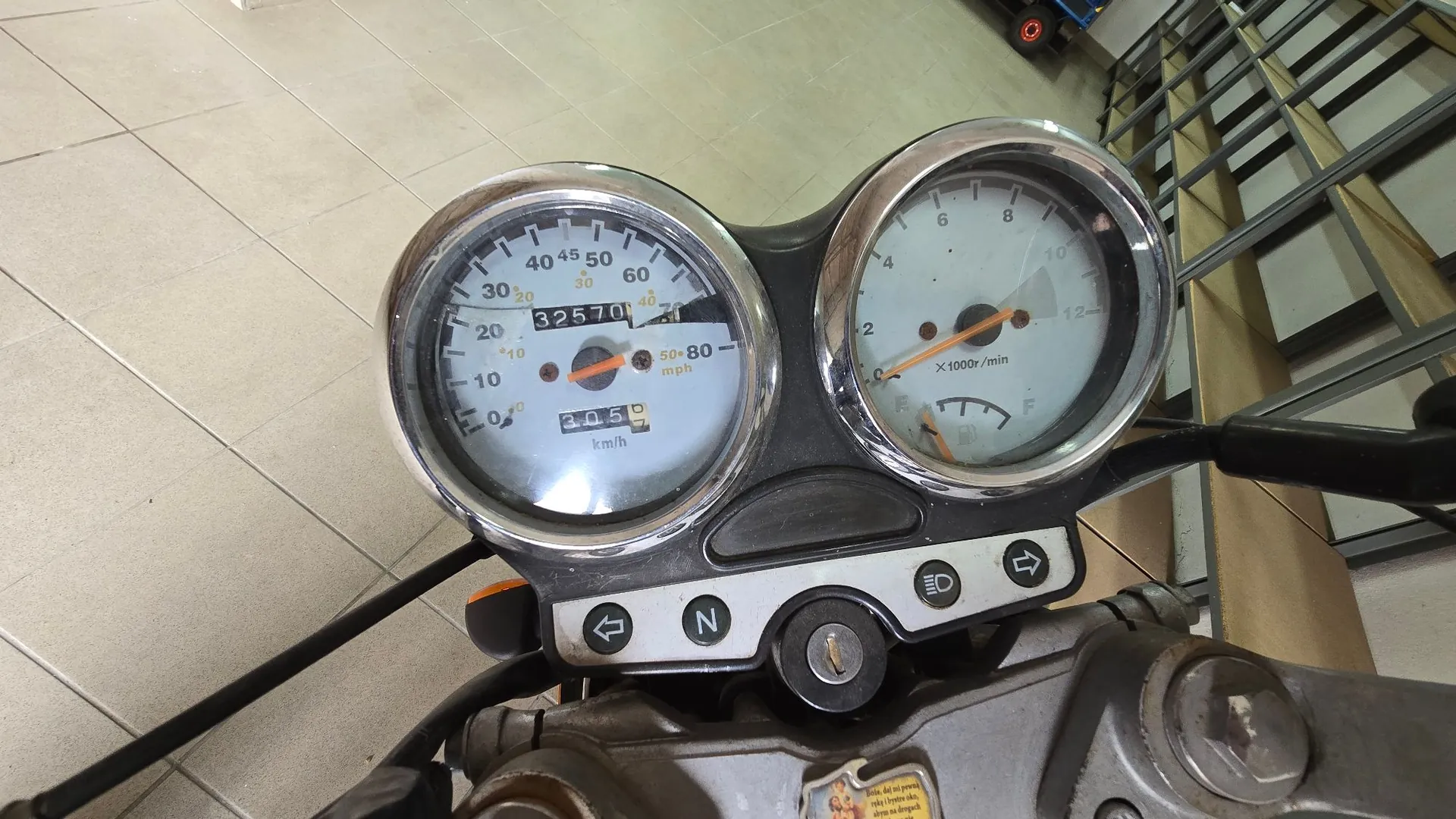 Zbliżenie na zestaw wskaźników motocyklowych. Po lewej stronie znajduje się prędkościomierz z dwiema skalami: zewnętrzną w km/h (od 0 do 100) i wewnętrzną w mph (od 0 do 60). Licznik przebiegu wskazuje wartość 32570. Po prawej stronie znajduje się obrotomierz z skalą od 0 do 12 (x1000 r/min) oraz wskaźnikiem poziomu paliwa (E-F). Poniżej zegarów widoczny jest panel z przyciskami, w tym wskaźnikiem neutralnego biegu (N) oraz przełącznikiem kierunkowskazów.
