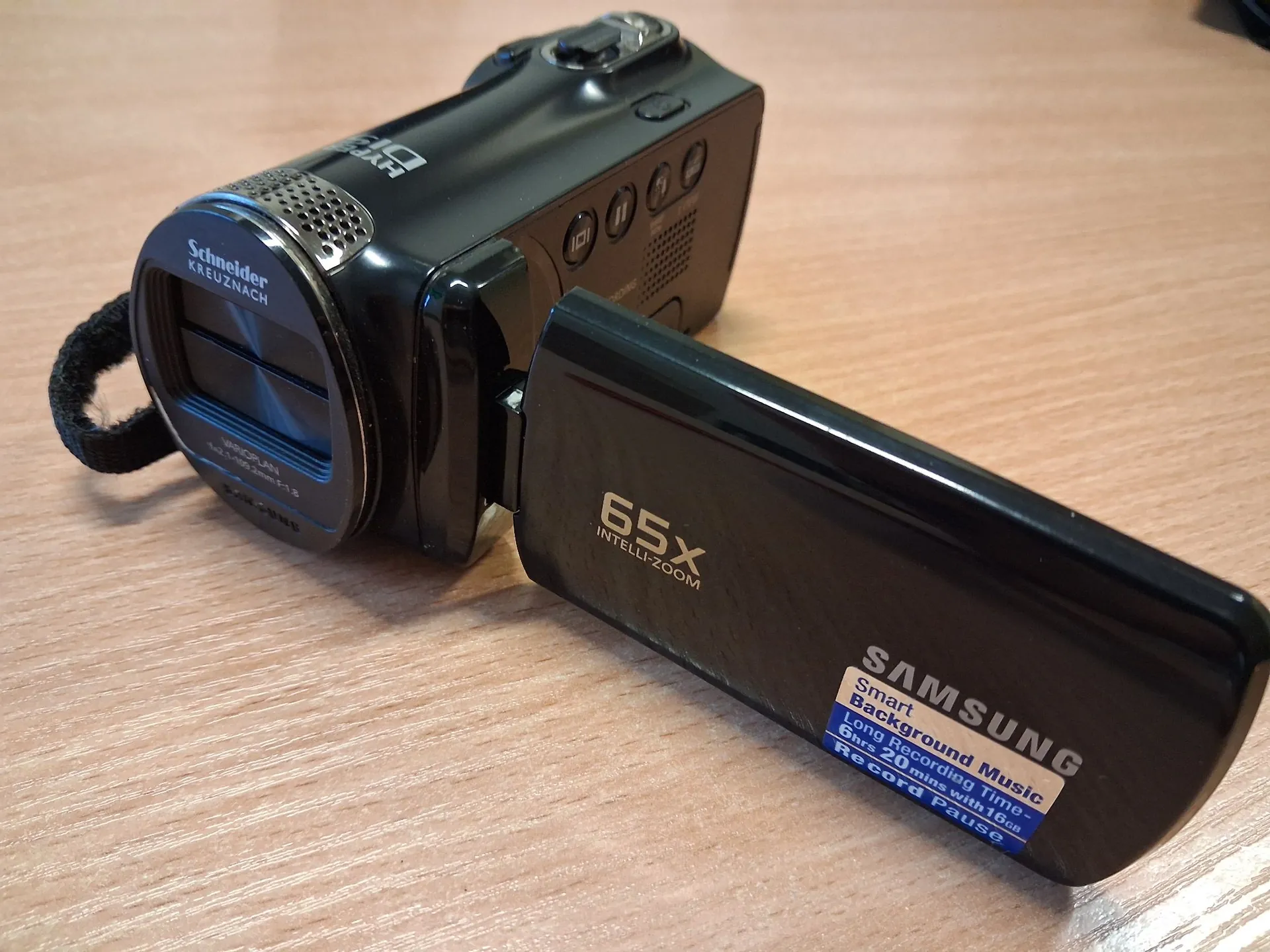 Czarna kamera wideo marki Samsung leżąca na drewnianym blacie. Na obiektywie widnieje napis 'Schneider KREUZNACH'. Wysunięty ekran LCD wyświetla napisy: '65X INTELLI-ZOOM' oraz naklejkę z tekstem: 'SAMSUNG Smart Background Music Long Recording time - 6hrs 20mins with 64GB Record Pause'.