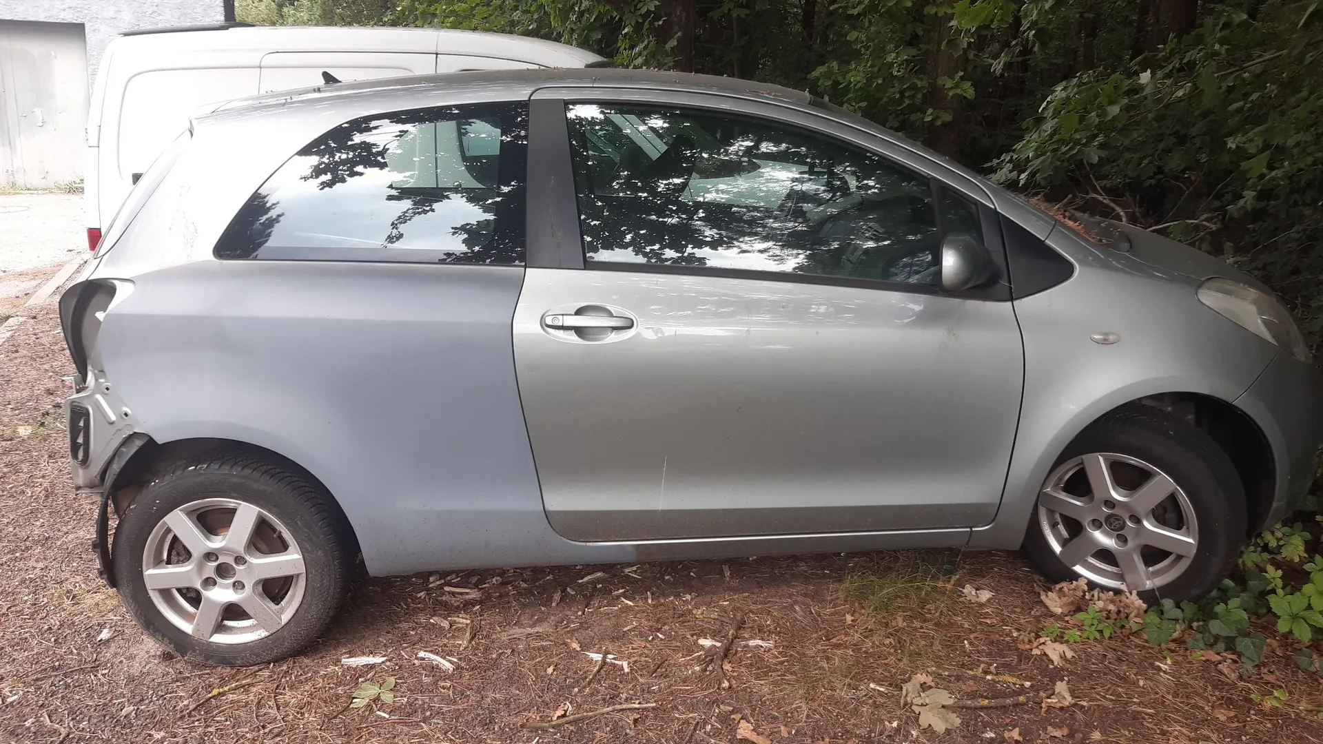 Srebrny samochód osobowy typu hatchback zaparkowany bokiem na leśnej ściółce. Pojazd ma widoczne uszkodzenia z tyłu, w tym brak tylnego zderzaka i wgniecenie błotnika. W tle widoczny jest biały furgon oraz gęste, zielone drzewa i krzewy.