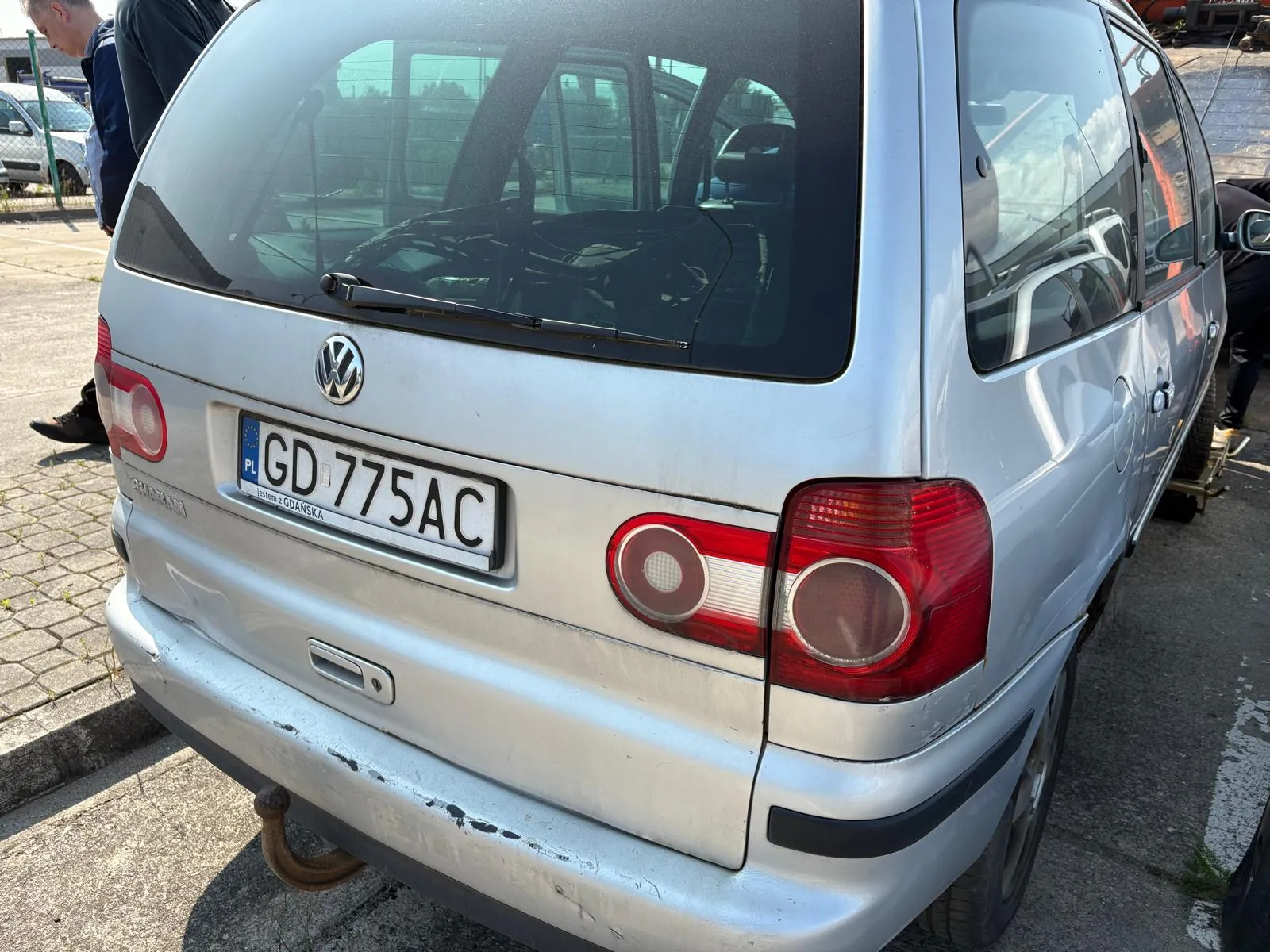 Srebrny samochód osobowy Volkswagen Sharan z tyłu. Widoczna tablica rejestracyjna PL GD 775AC z napisem "Jestem z GDANSKA" poniżej. Na zderzaku ślady uszkodzeń i rdzewiejący hak holowniczy. Czerwone światła tylne, logo VW na bagażniku. Po lewej stronie osoba w ciemnej odzieży, na tle brukowany plac i częściowo widoczny inny pojazd. W tle siatka ogrodzeniowa i drzewa.