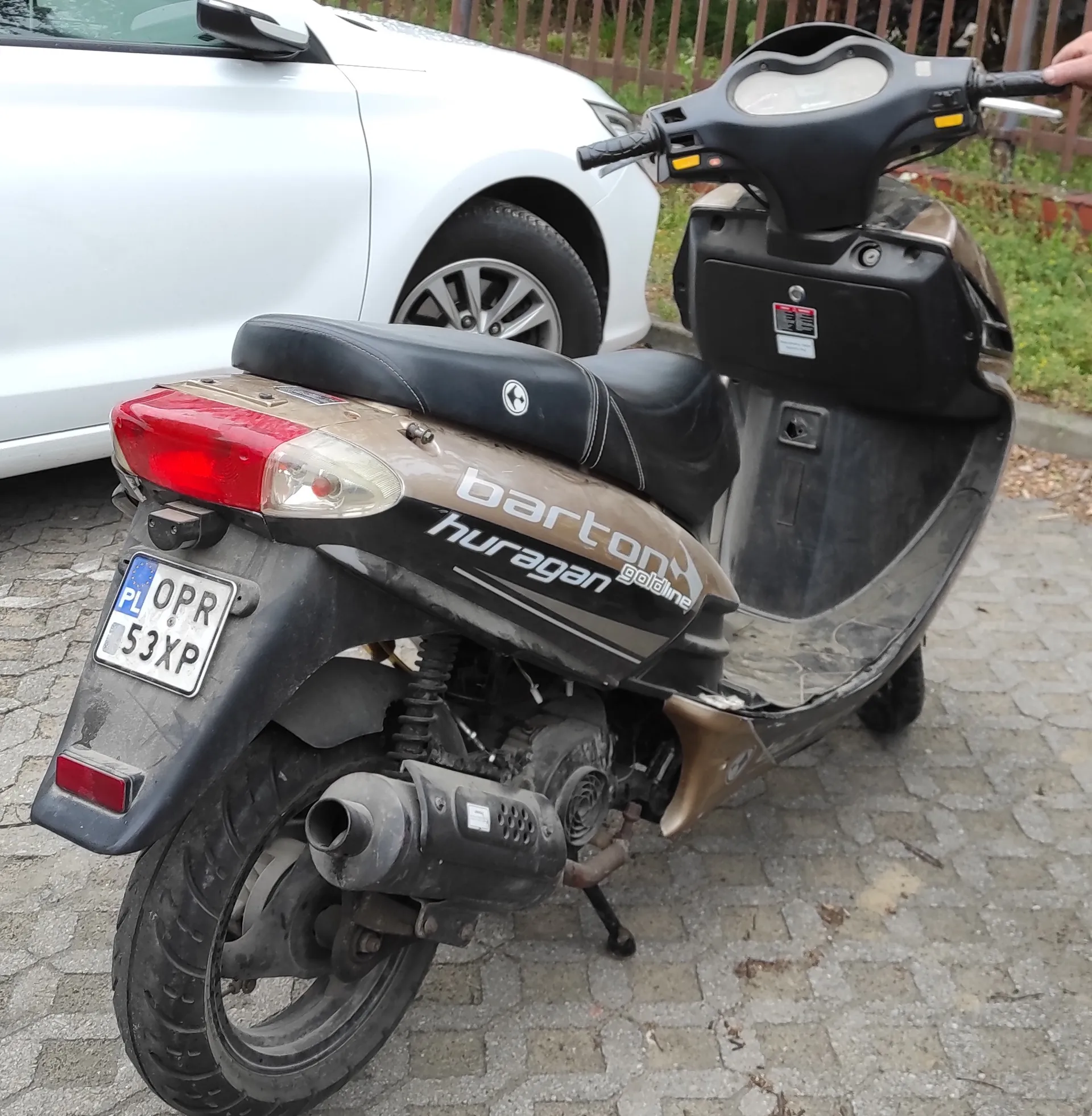 Widok z tyłu i z boku brązowego skutera marki Barton, model Huragan Gold Line. Skuter posiada czarną kanapę, czarną owiewkę przednią oraz czarną tylną błotnik. Na tylnym błotniku zamontowana jest tablica rejestracyjna o numerze OPR 53XP z oznaczeniem PL. W tle widoczny jest fragment białego samochodu oraz kostka brukowa na ziemi.