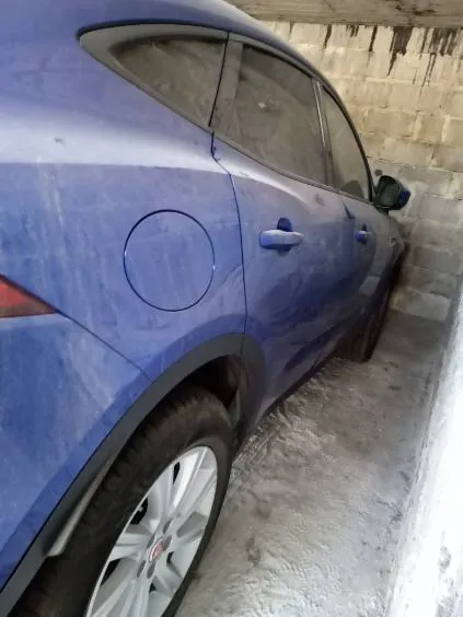 Niebieski SUV zaparkowany w wąskim przejściu między betonowymi ścianami. Widoczna tylna część pojazdu z kołem, pokrywą zbiornika paliwa oraz fragmentem bocznej szyby. Ściany betonowe mają plamy i przebarwienia, podłoga również jest betonowa z widocznymi zabrudzeniami.