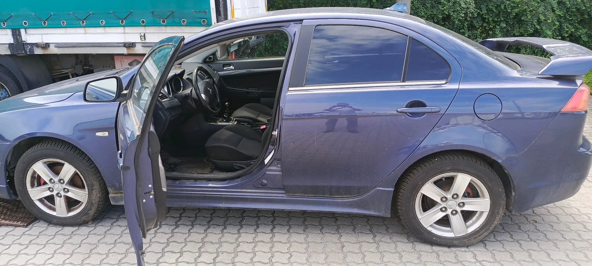 Niebieski samochód osobowy typu sedan zaparkowany na kostce brukowej. Przednie lewe drzwi są otwarte, ukazując wnętrze pojazdu z czarnymi fotelami i kierownicą. Samochód posiada srebrne felgi aluminiowe oraz spojler na klapie bagażnika. W tle widoczny jest fragment białego pojazdu ciężarowego z zielną nadbudową oraz zielona roślinność.