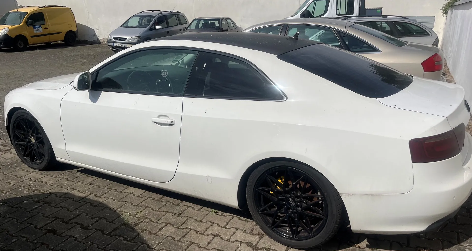 Biały samochód osobowy typu coupe z czarnymi felgami, zaparkowany na kostce brukowej. W tle widoczne są inne zaparkowane pojazdy: żółty furgon oraz srebrne samochody osobowe.