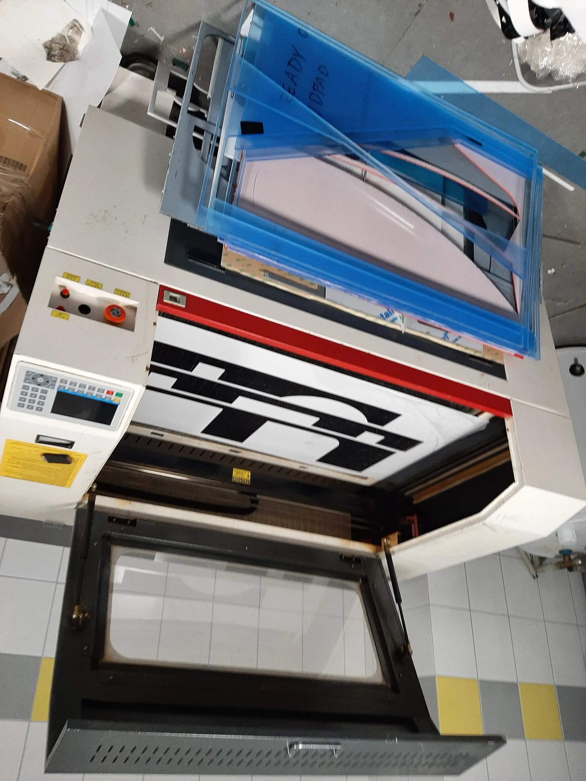 Przemysłowa maszyna o białym kolorze z otwartą czarną drzwiami z szybą. Na górze maszyny znajduje się panel sterowania z przyciskami, ekranem i czerwonymi oraz pomarańczowymi guzikami. Wewnątrz maszyny widoczny czarno-biały wzór. Po prawej stronie stos niebieskich płyt z napisem "READY" i częściowo widocznym "DRAD". Tło stanowi biała płytkowa ściana z żółtymi i szarymi pasami oraz betonowa podłoga. W tle widoczne są pudełko kartonowe i inne elementy warsztatu.