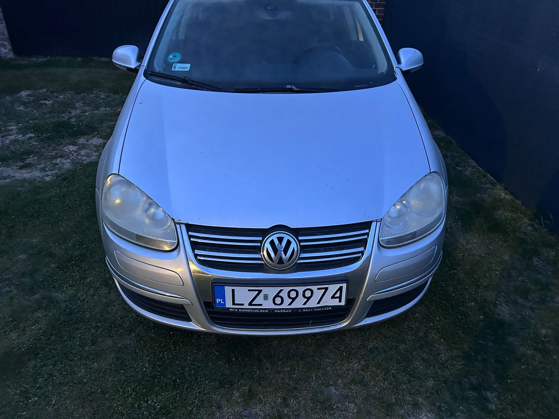 Srebrny samochód marki Volkswagen widziany od przodu, zaparkowany na trawie. Na masce widoczne jest logo marki, a poniżej tablica rejestracyjna o numerze LZ 69974 z oznaczeniem PL. Pod tablicą znajduje się napis: RFZ BURGHOLZER - PASSAU - 0651-5067234.