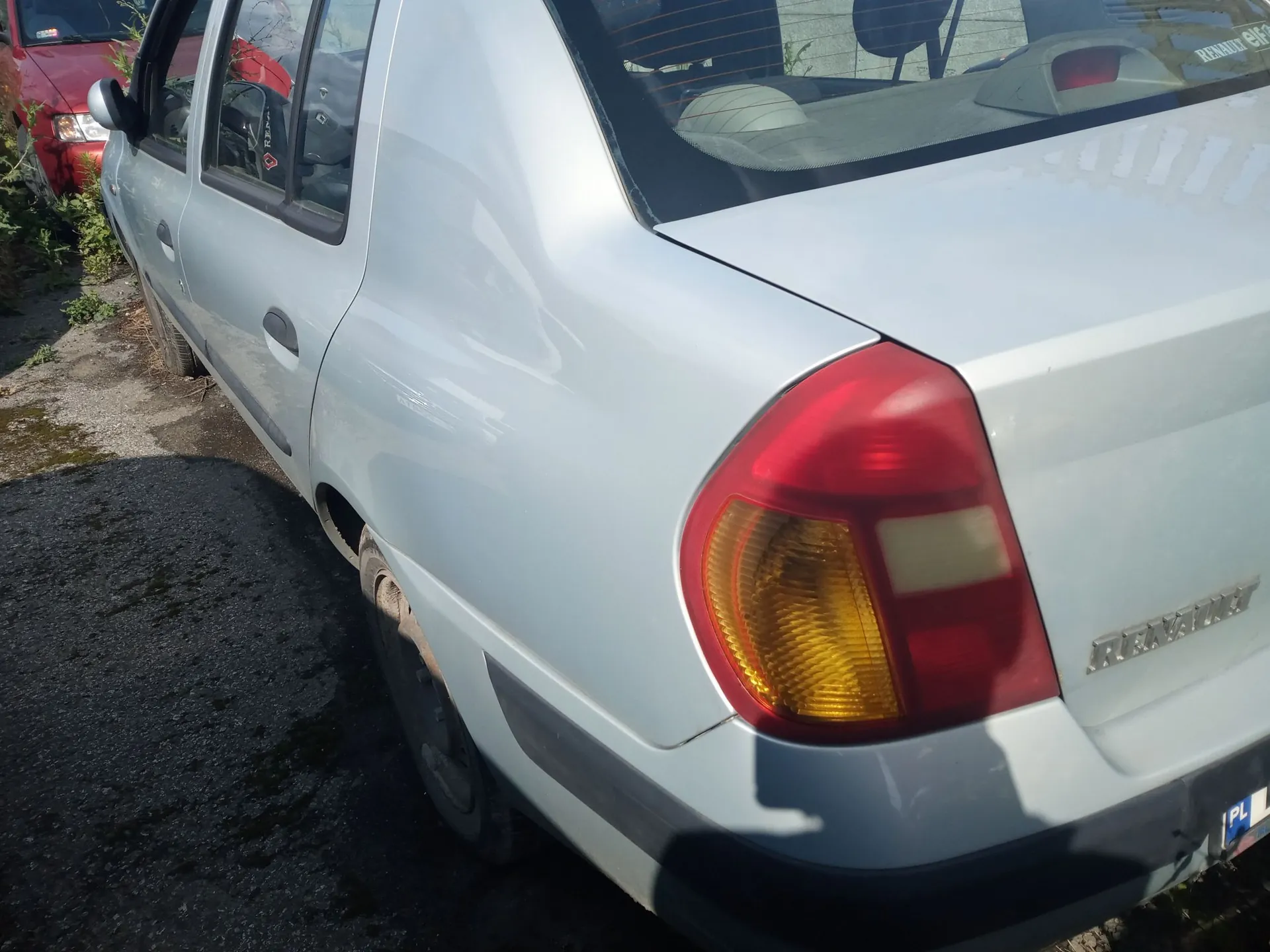Srebrny samochód marki Renault widziany z tyłu z boku. Widoczna jest tylna prawa lampa, tylny zderzak oraz fragment bocznej części nadwozia i tylnego koła. W tle widoczny jest fragment czerwonego samochodu.